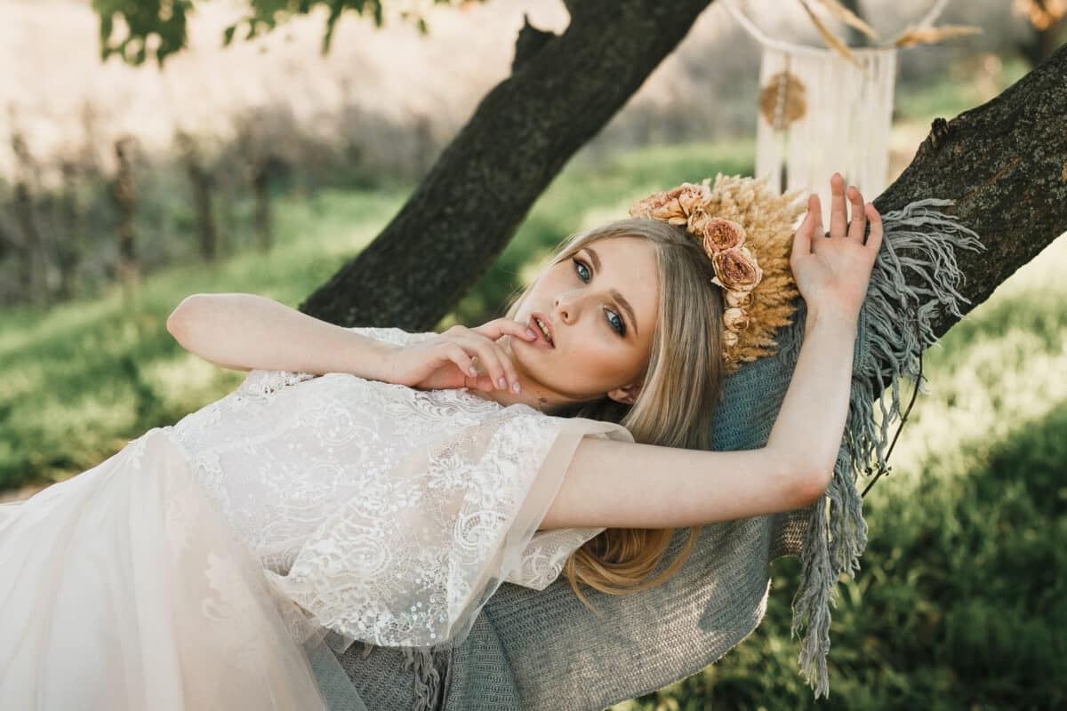 a pretty lady in white dress with a crown of dry grass on her head lying on a tree trunk in the woods