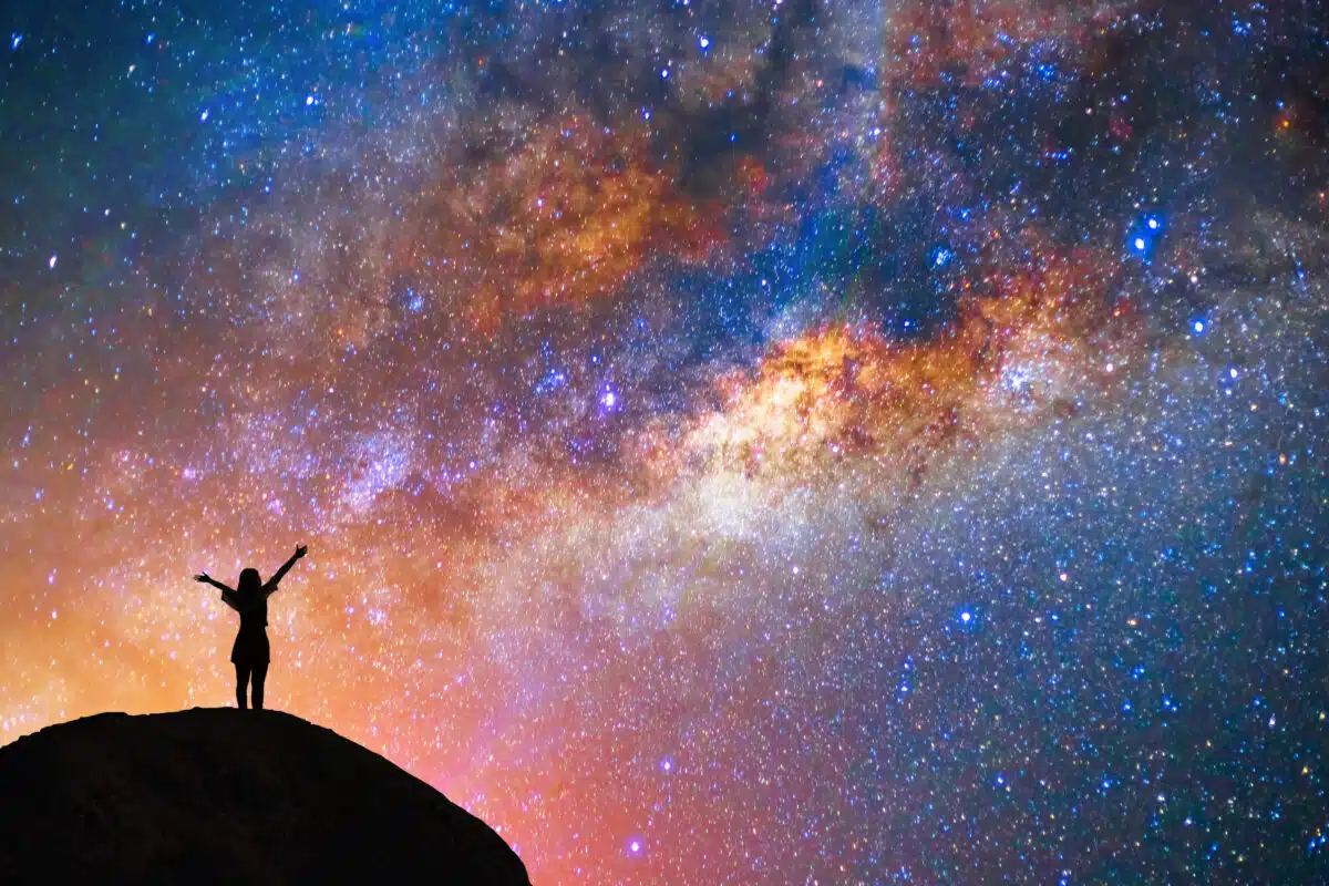 a happy woman on the mountain raising her arms in the beautiful milky way above her