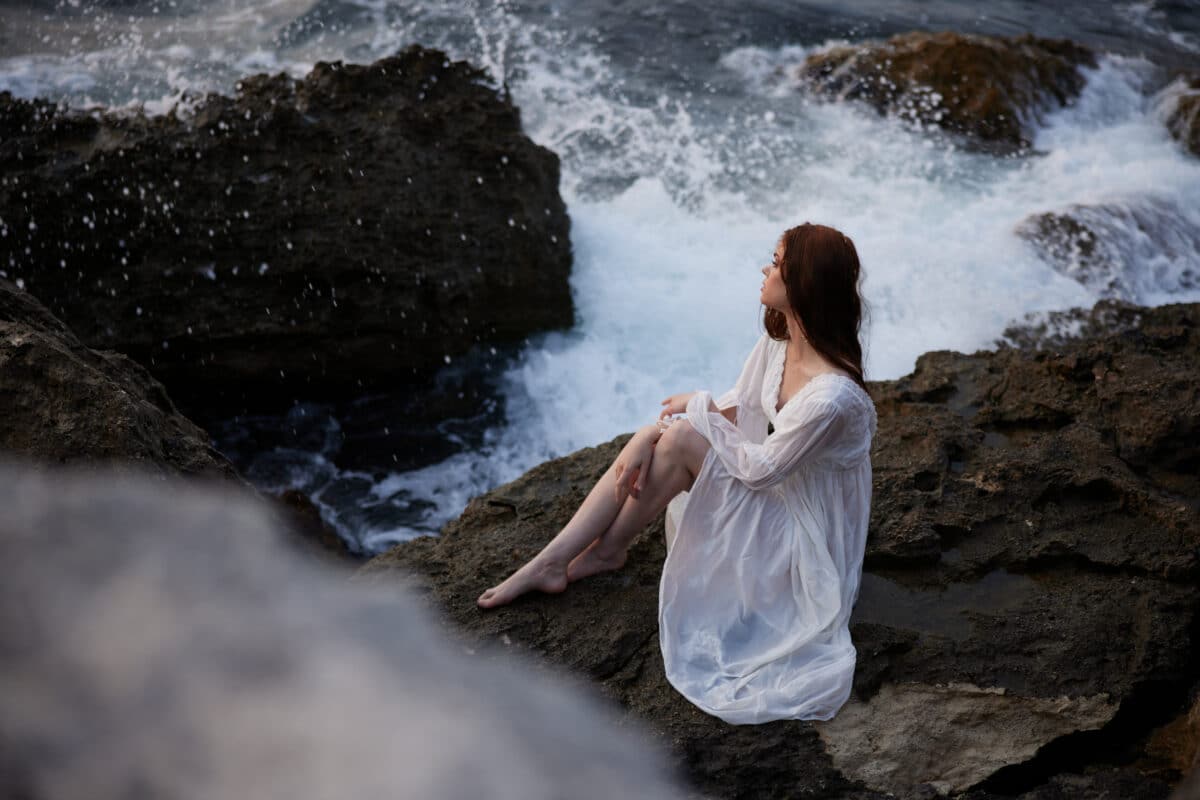 woman in white dress sits on stones by the ocean