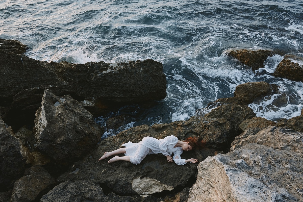 red haired woman with long hair in a white dress lying on a stone by the ocean