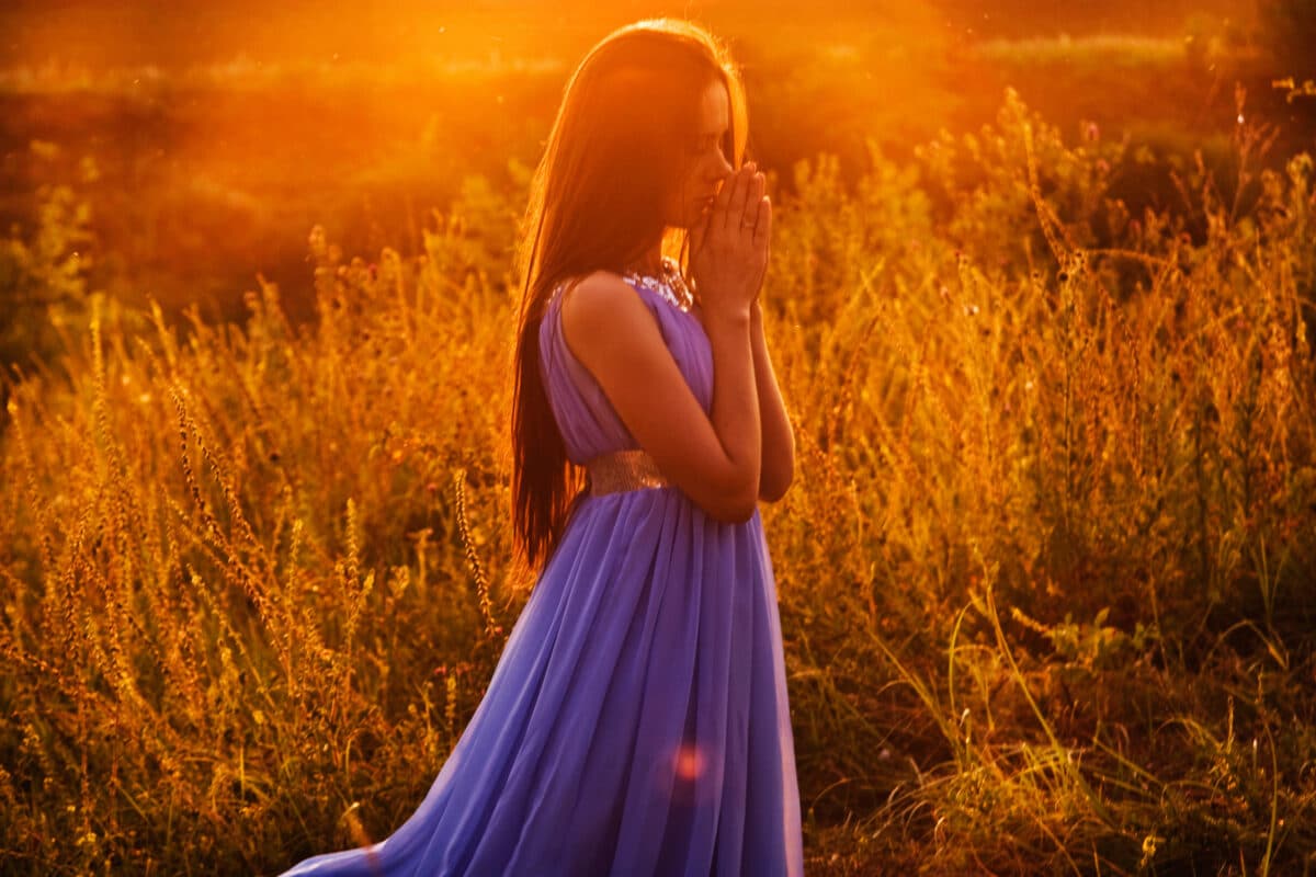 Beautiful girl kneeling and praying in the field at sunset