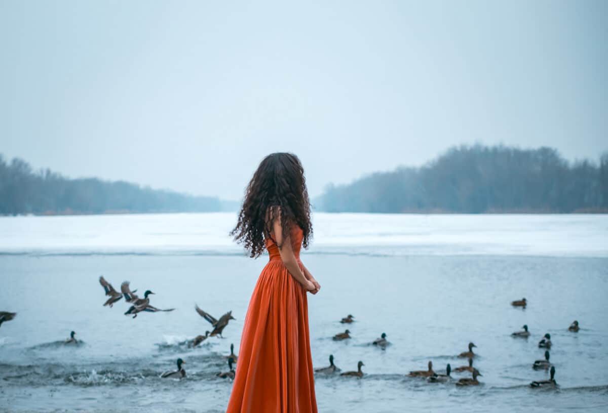 Beautiful young brunette woman standing by the cold lake with birds in the winter forest