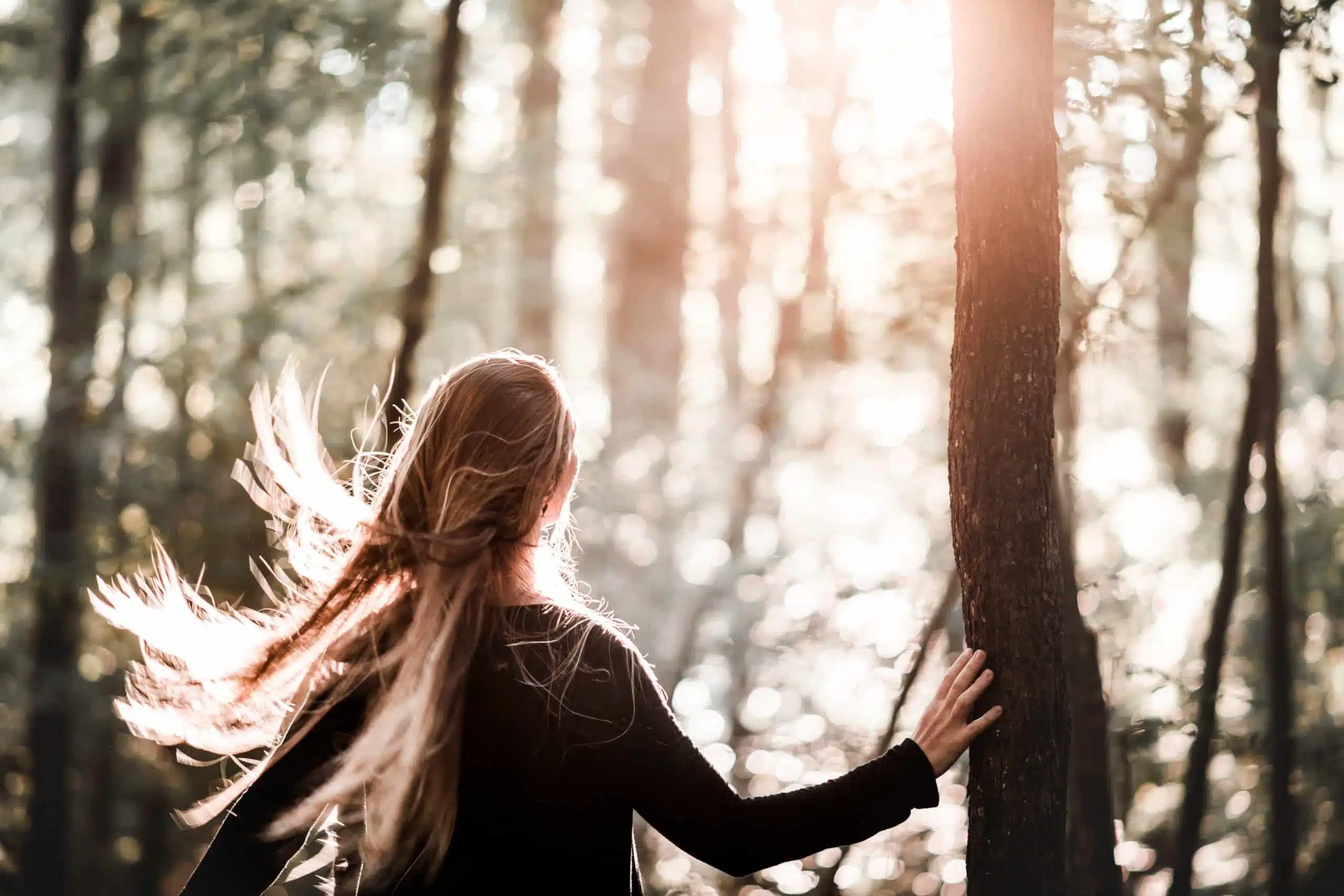 Woman standing in the woods with sunlight, hair blown by the wind.