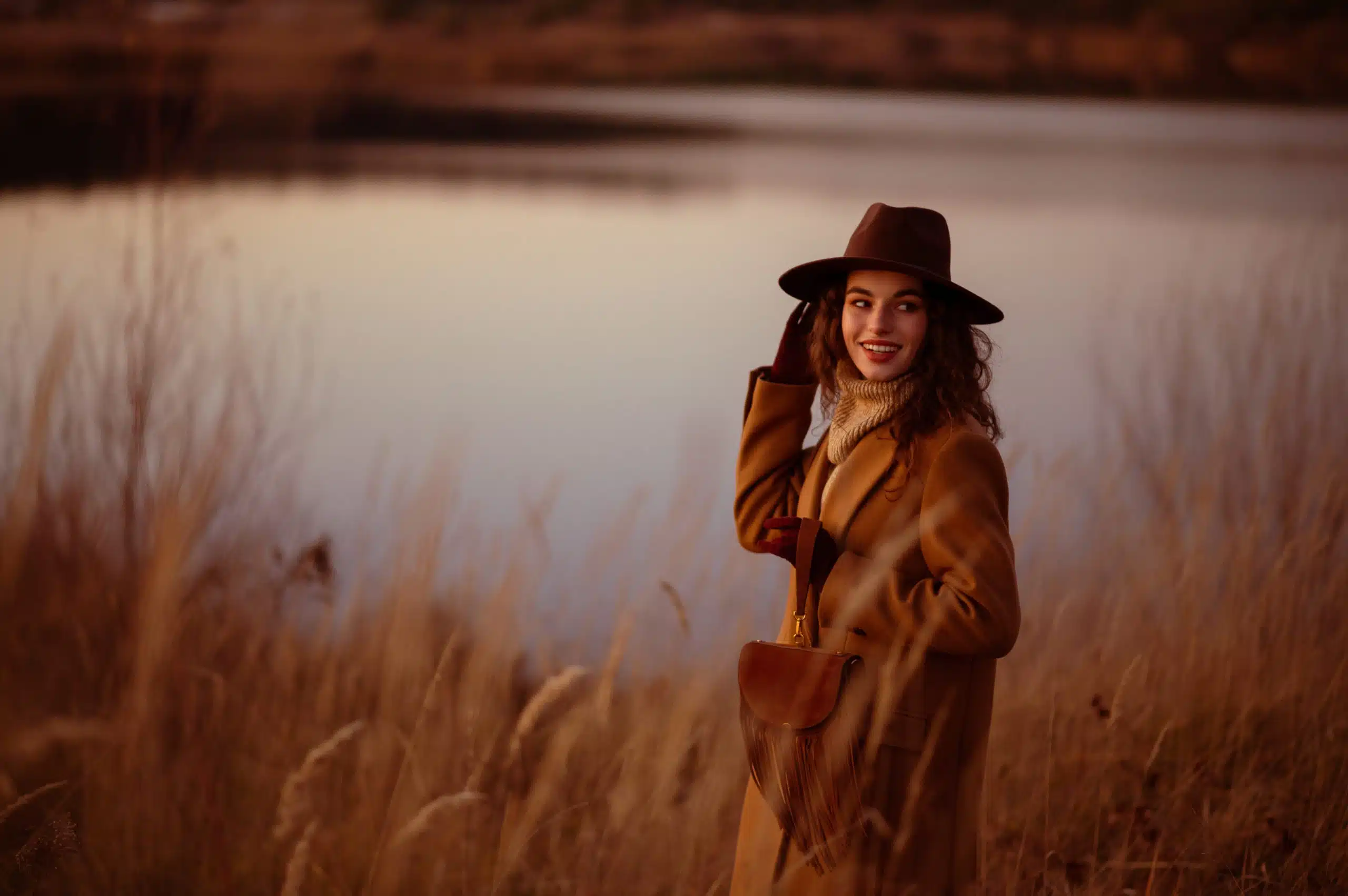 Standing in the grasses by the lake, a happy smiling elegant woman wearing a brown hat and coat.