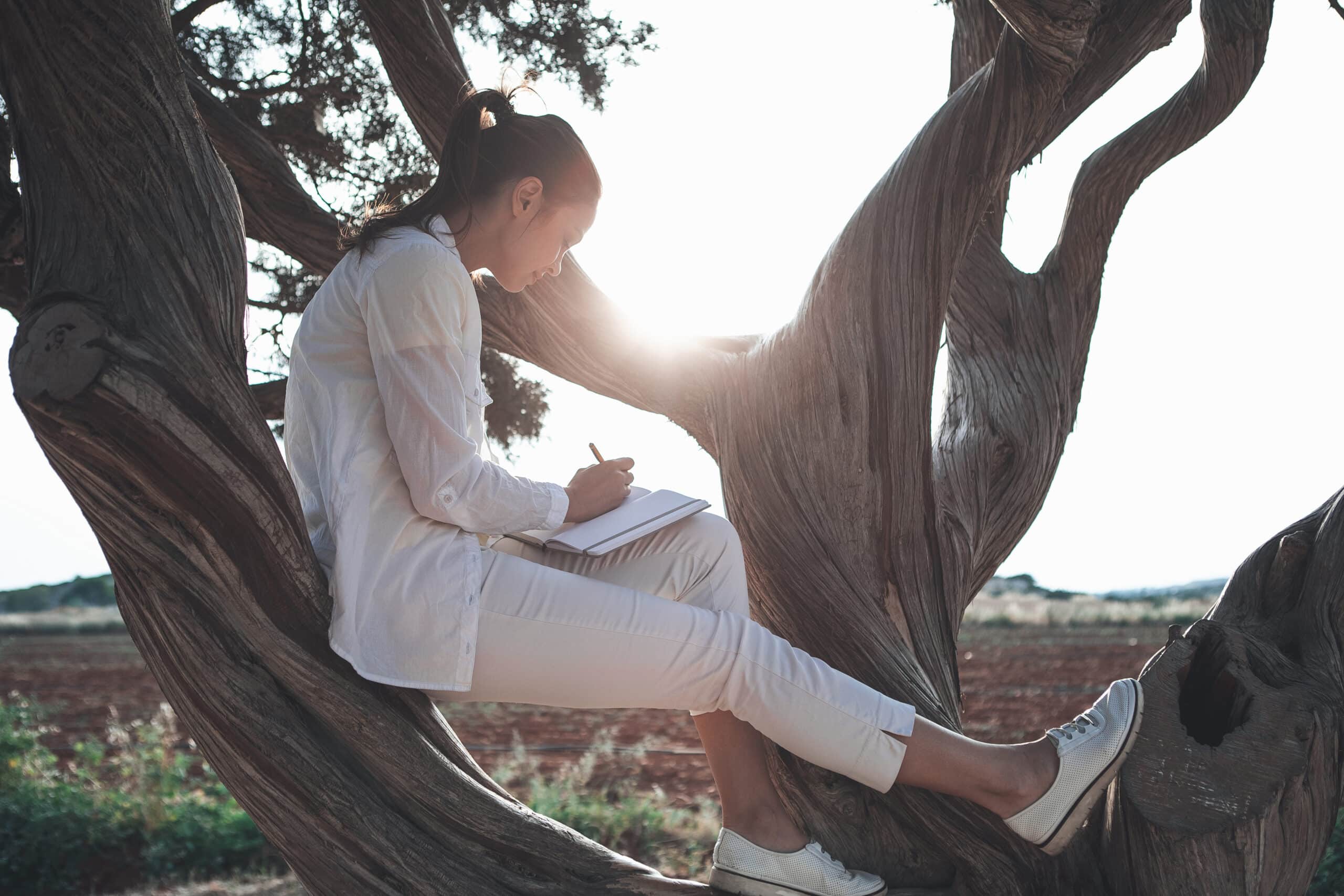 Woman in white writing while sitting on a low-lying tree.