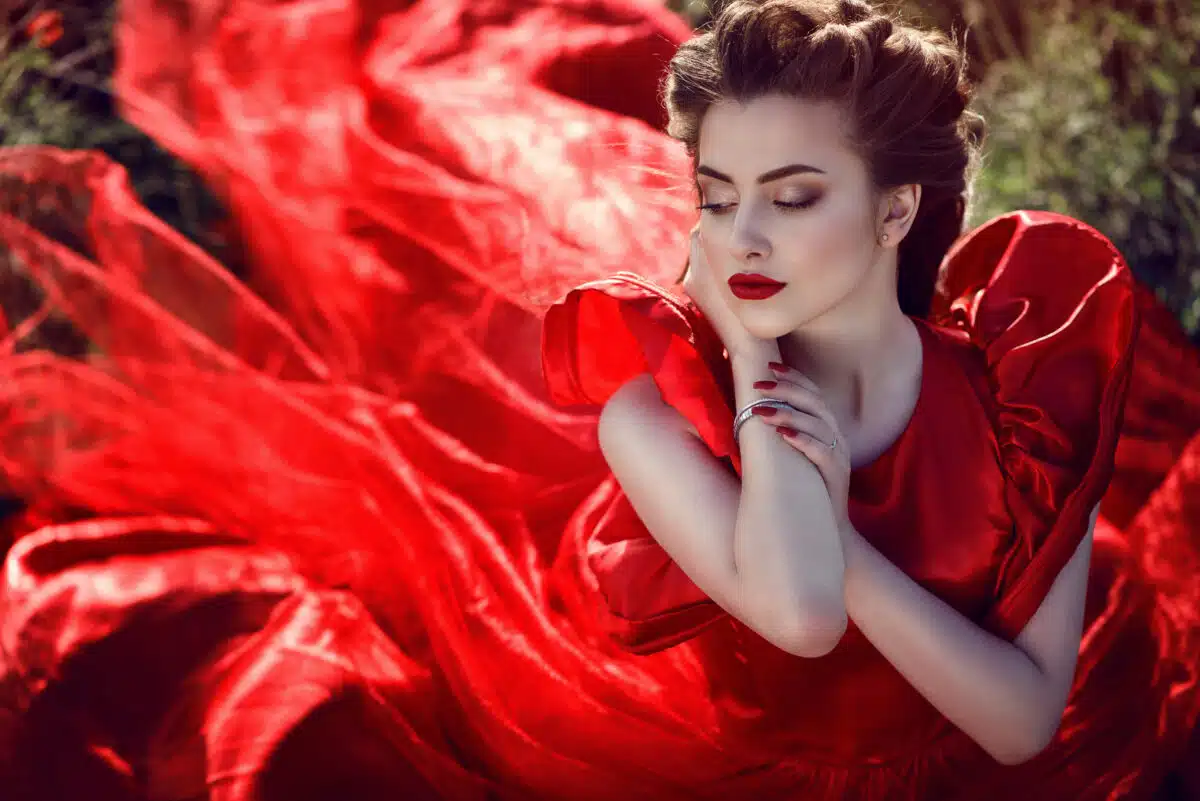 Beautiful young lady with perfect make up and plaited hairstyle wearing luxurious silk red ballroom dress sitting in the poppy field. The wind in her hair. The tail of her dress flying behind