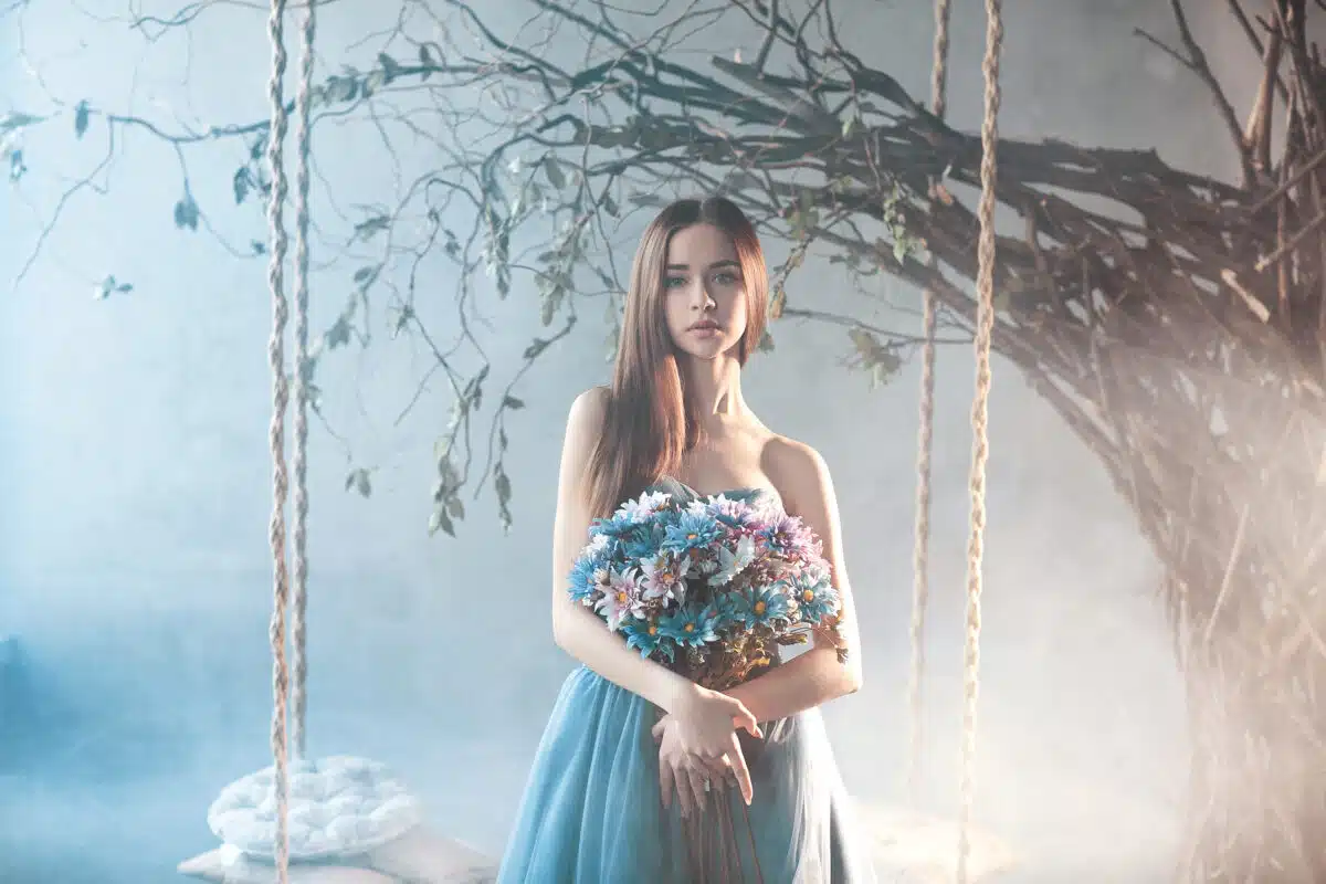 Beautiful gorgeous calm woman in long chiffon blue dress holding big bouquet of flowers standing in the forest near the tree with swing.