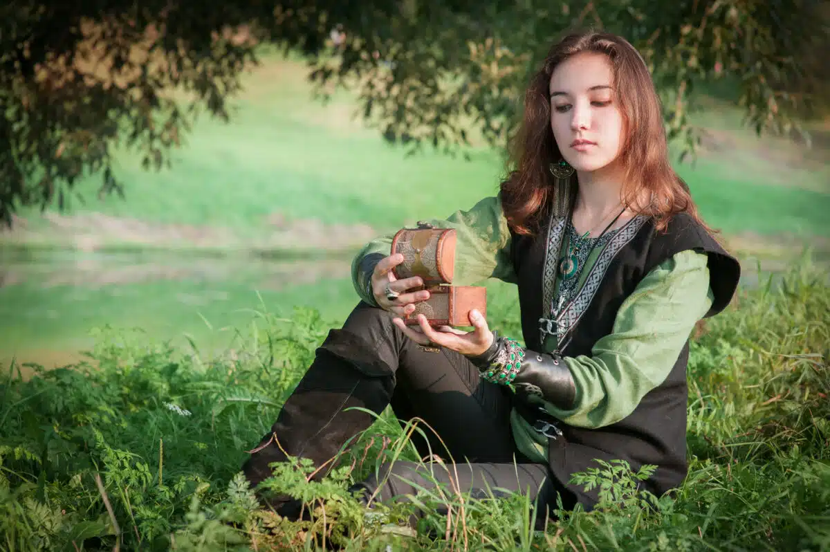 Young beautiful woman wearing man’s medieval style dress holding old casket outdoor.