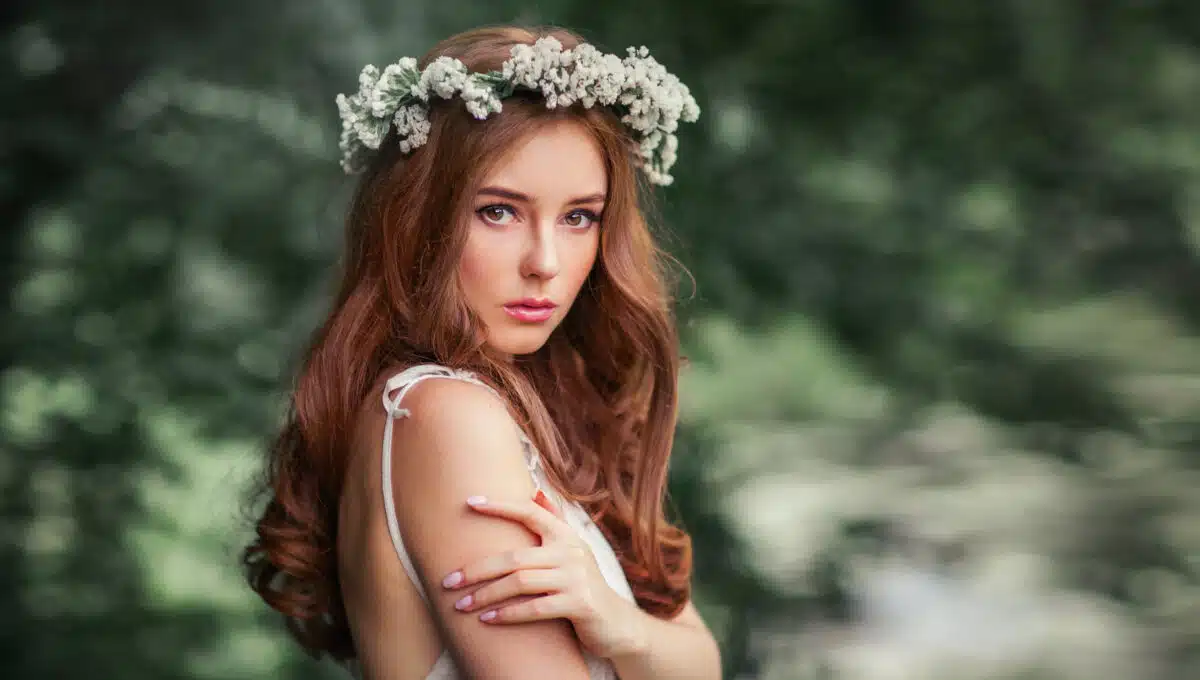 Beautiful red haired girl in white vintage dress and wreath of flowers standing under the tree and looking at camera. Fairytale story.