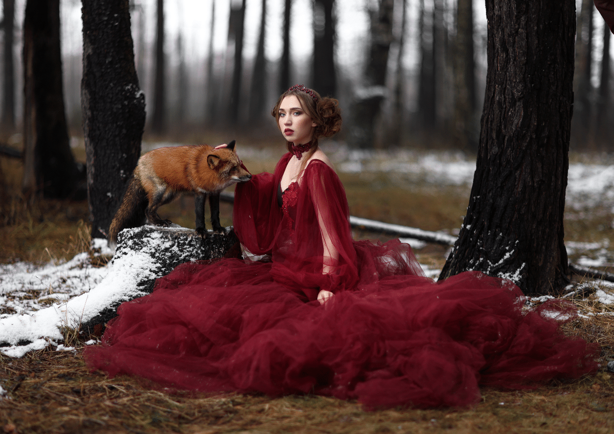 beautiful young woman in long red dress sitting near fox in the forest
