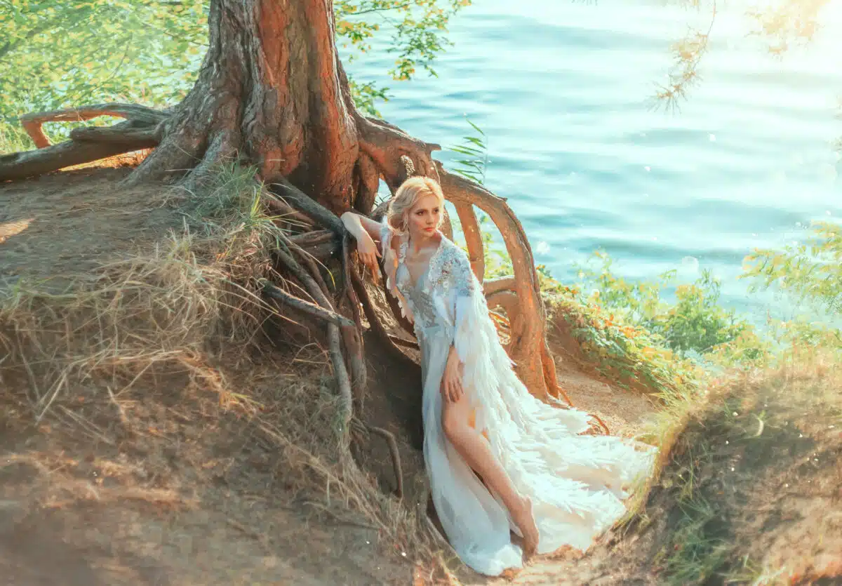 Luxurious blonde woman in a white dress with a long royal cape with feathers. Queen Swan stands near a tree with huge roots on the river coast. Background summer nature landscape, warm green colors.