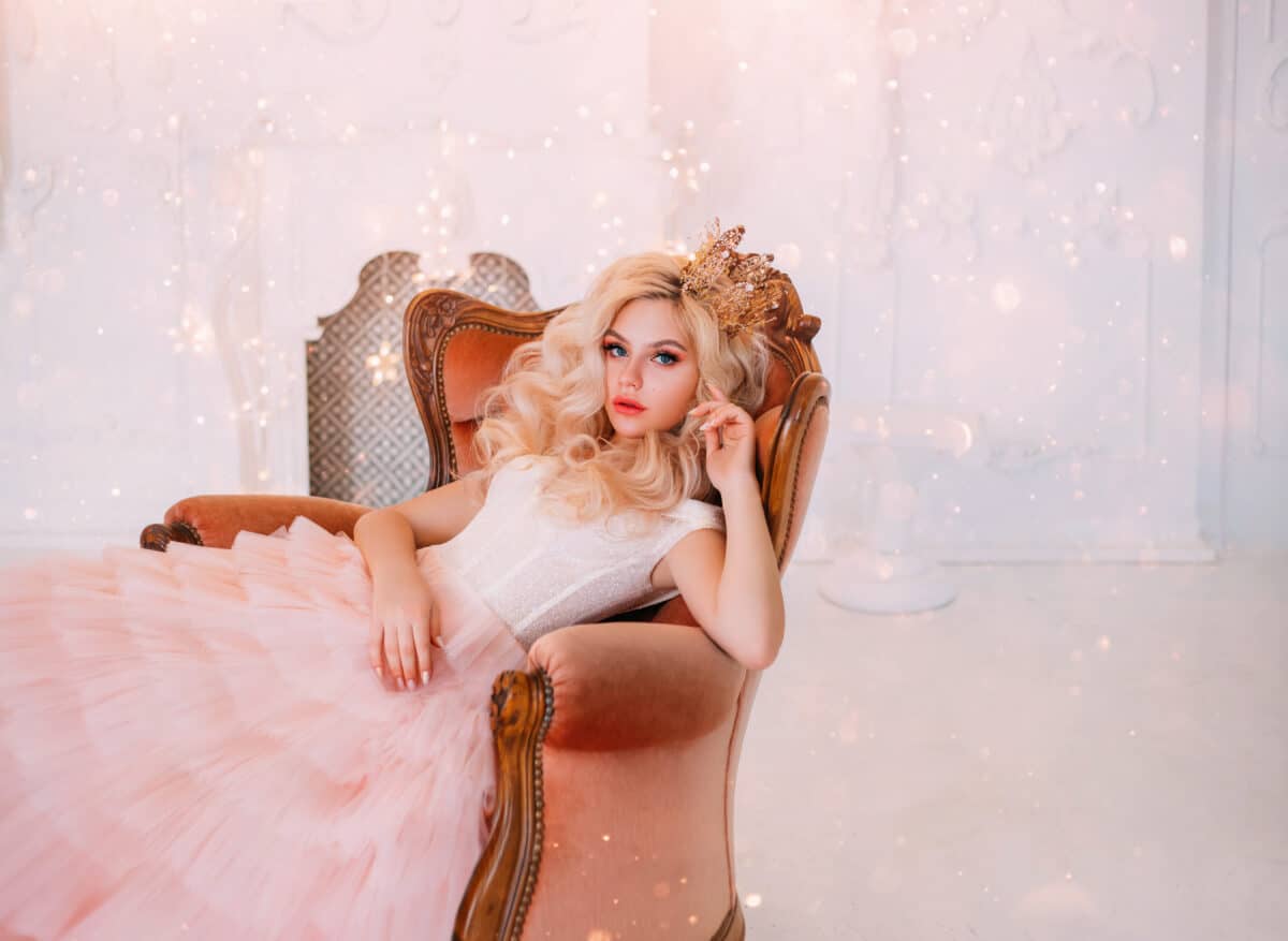 Young beautiful queen woman resting in medieval armchair. beauty face. New Year garland scenery glare sparks bright. Backdrop white classic room interior. Hairstyle princess girl blonde long wavy hair