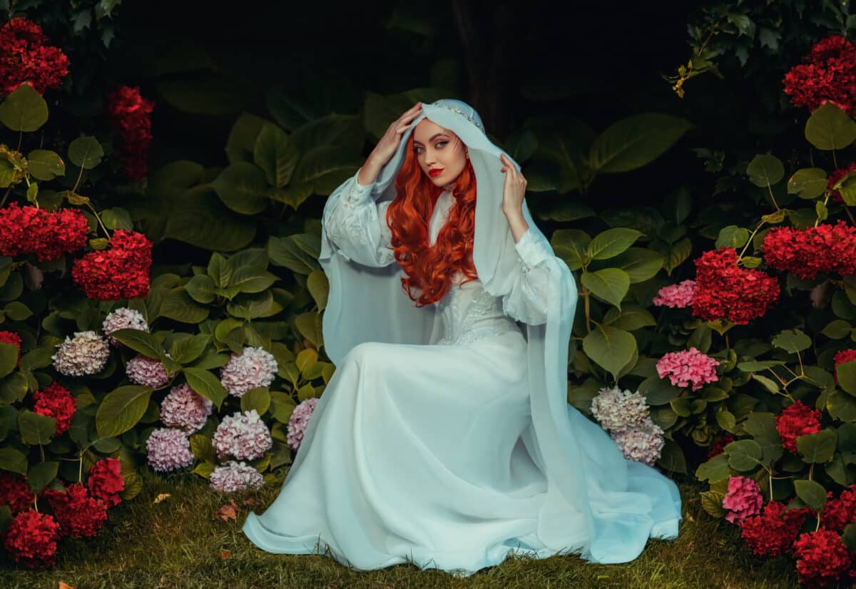 Fantasy woman in white vintage dress head covered with veil. Red-haired girl queen. Medieval princess bride sits in garden green trees bushes hydrangea flowers. Mystical image sexy lady gothic vampire