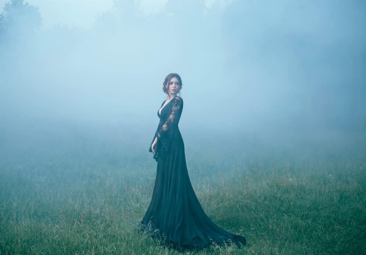 a girl in a black long dress walking along ia a clearing in thick fog. scared, beautiful, witch ia going to the forest, looking for a house. art photo. halloween costume. model. dark. mystic. evil.