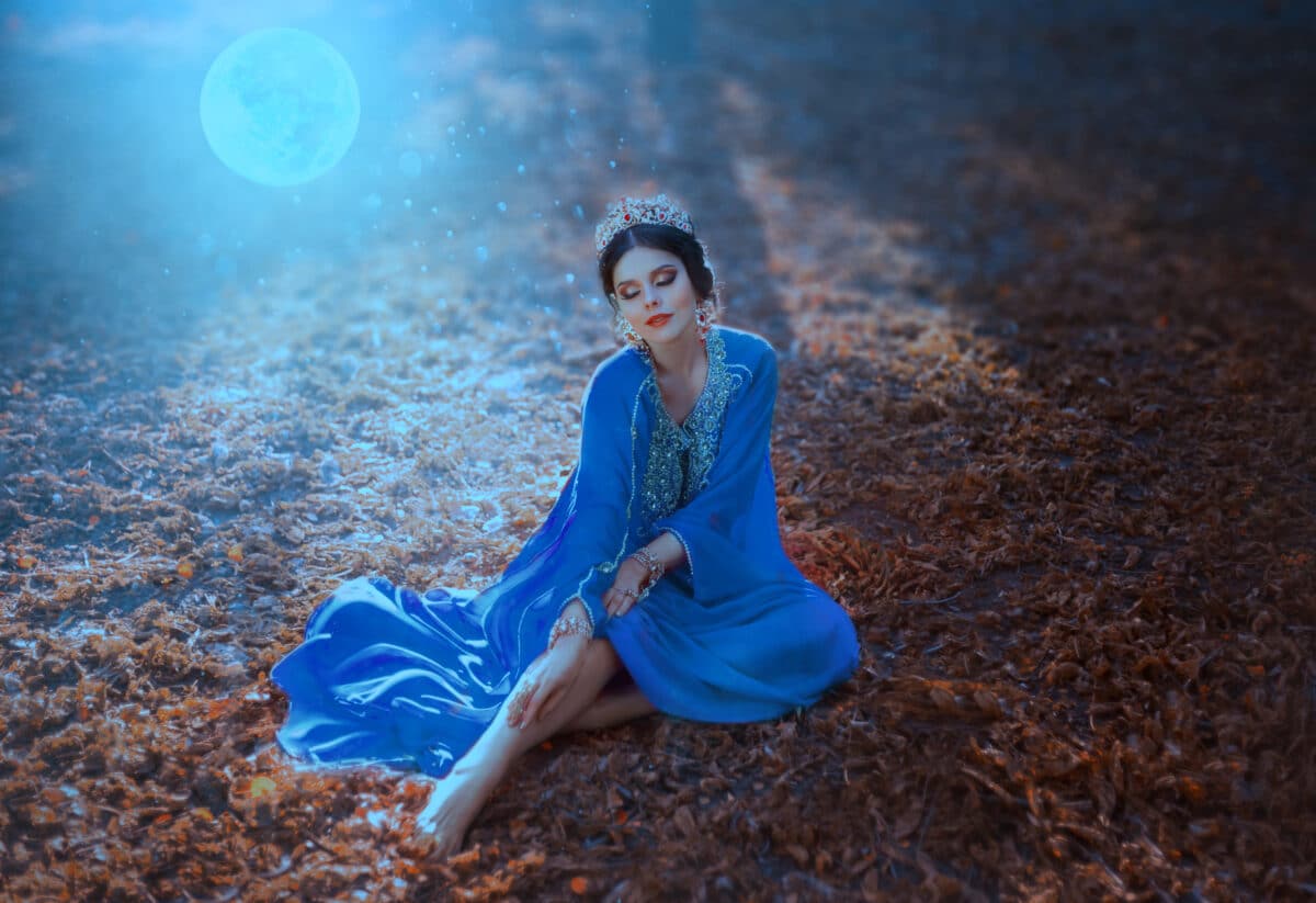 Young woman with the moon. Night autumn landscape. The Eastern Queen in a blue dress sits on the ground covered with fallen leaves in the moonlight. Tales of Shikhirizada 1001 nights. Art photography