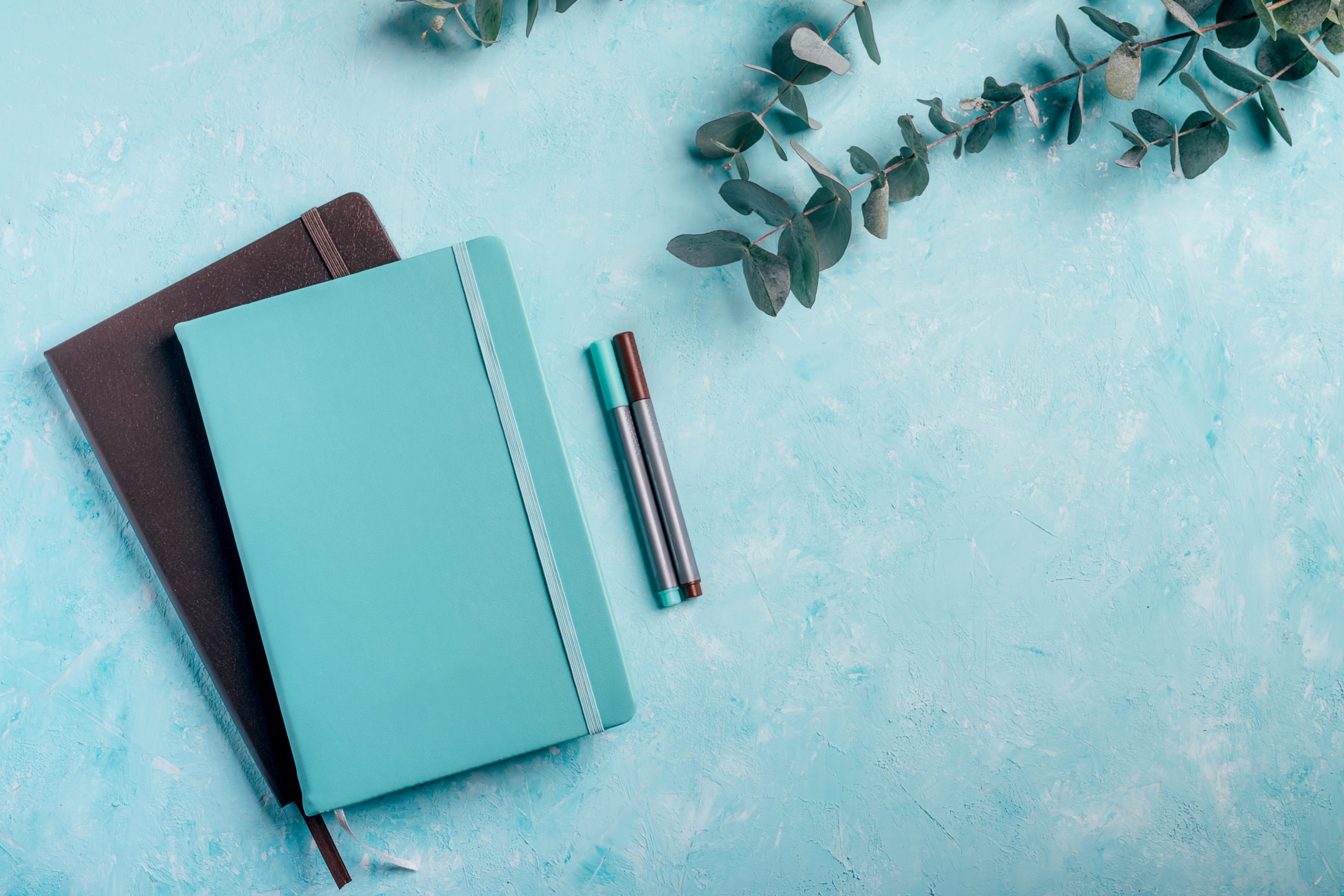 blue and brown notebooks, markers with green eucalyptus plants on turquoise rustic desk background