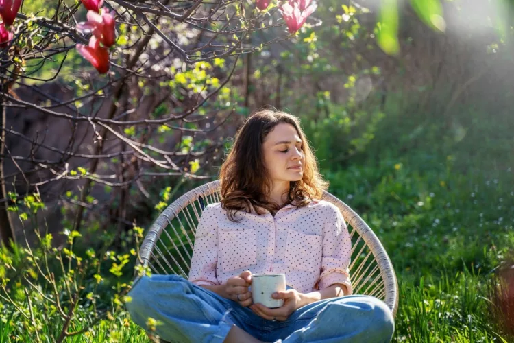 Young beautiful woman enjoying spring time while sitting in blooming garden of her home with a cup of tea.
