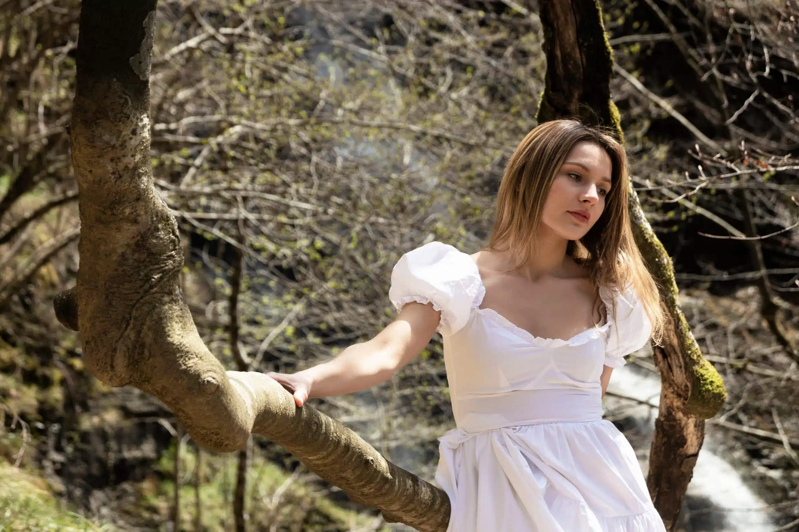 blonde woman in a white dress sitting on a tree in the forest