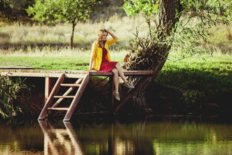 carefree young woman sitting on a wooden river bridge with a book nearby and enjoying nature.