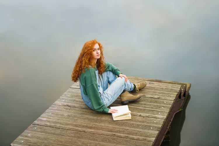 relaxed redhead woman reading a book on wooden pontoon.