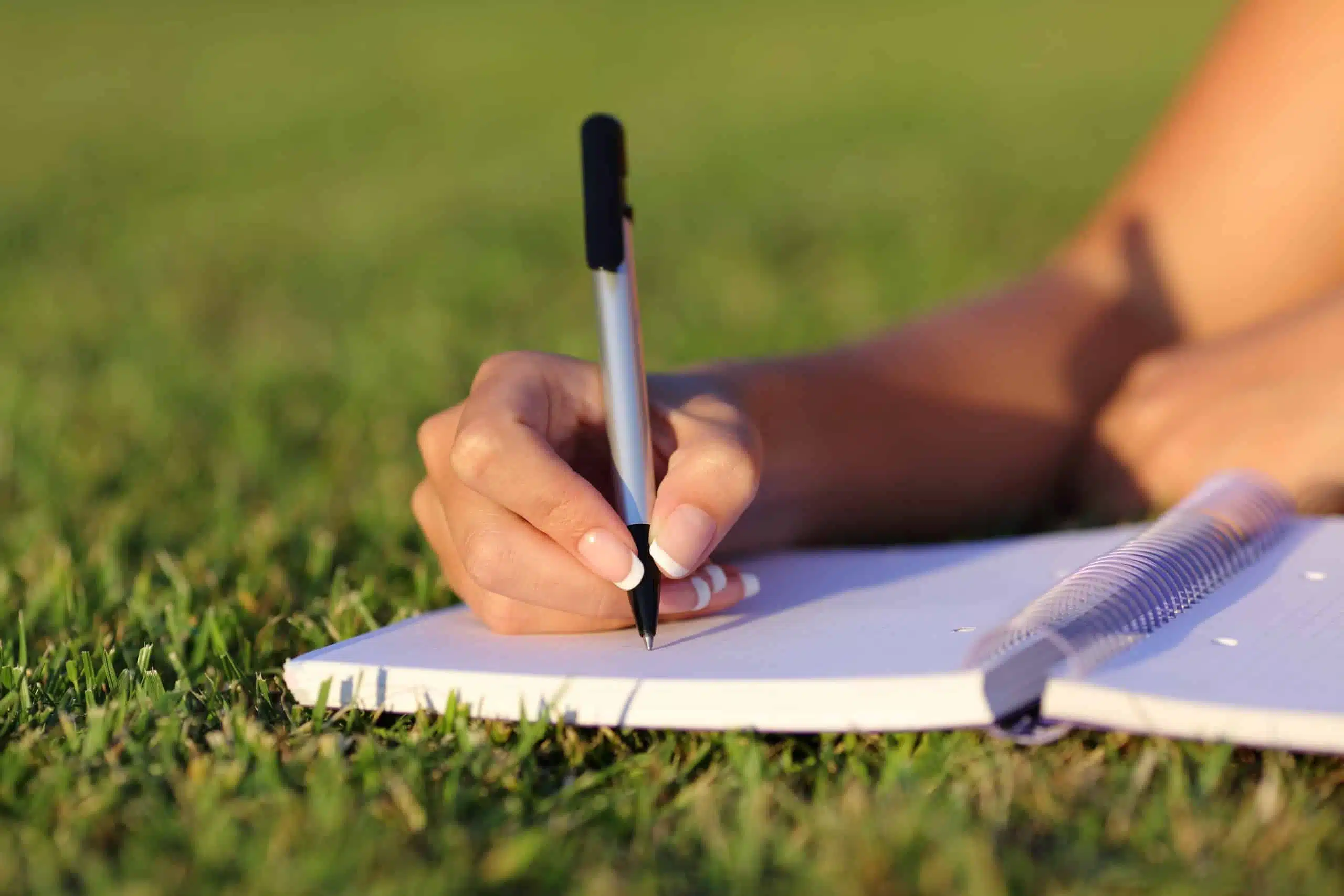 A female hand writing on a notebook outdoor on the grass.