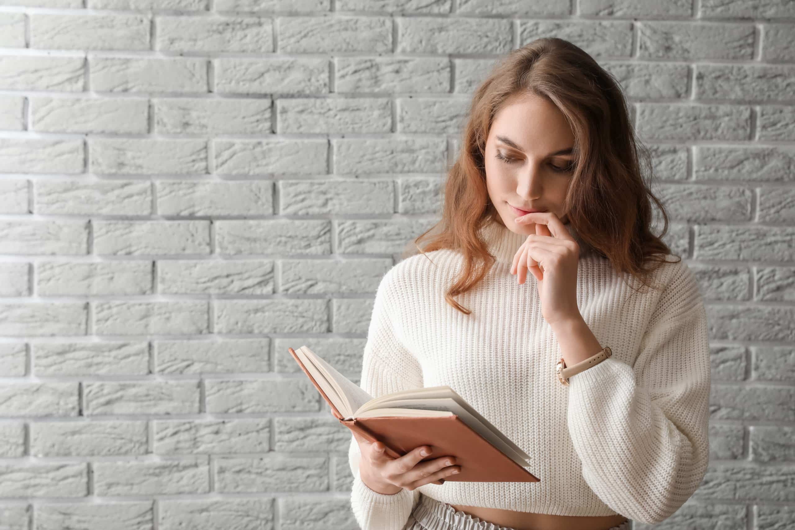Pensive woman reading book near grey brick wall.