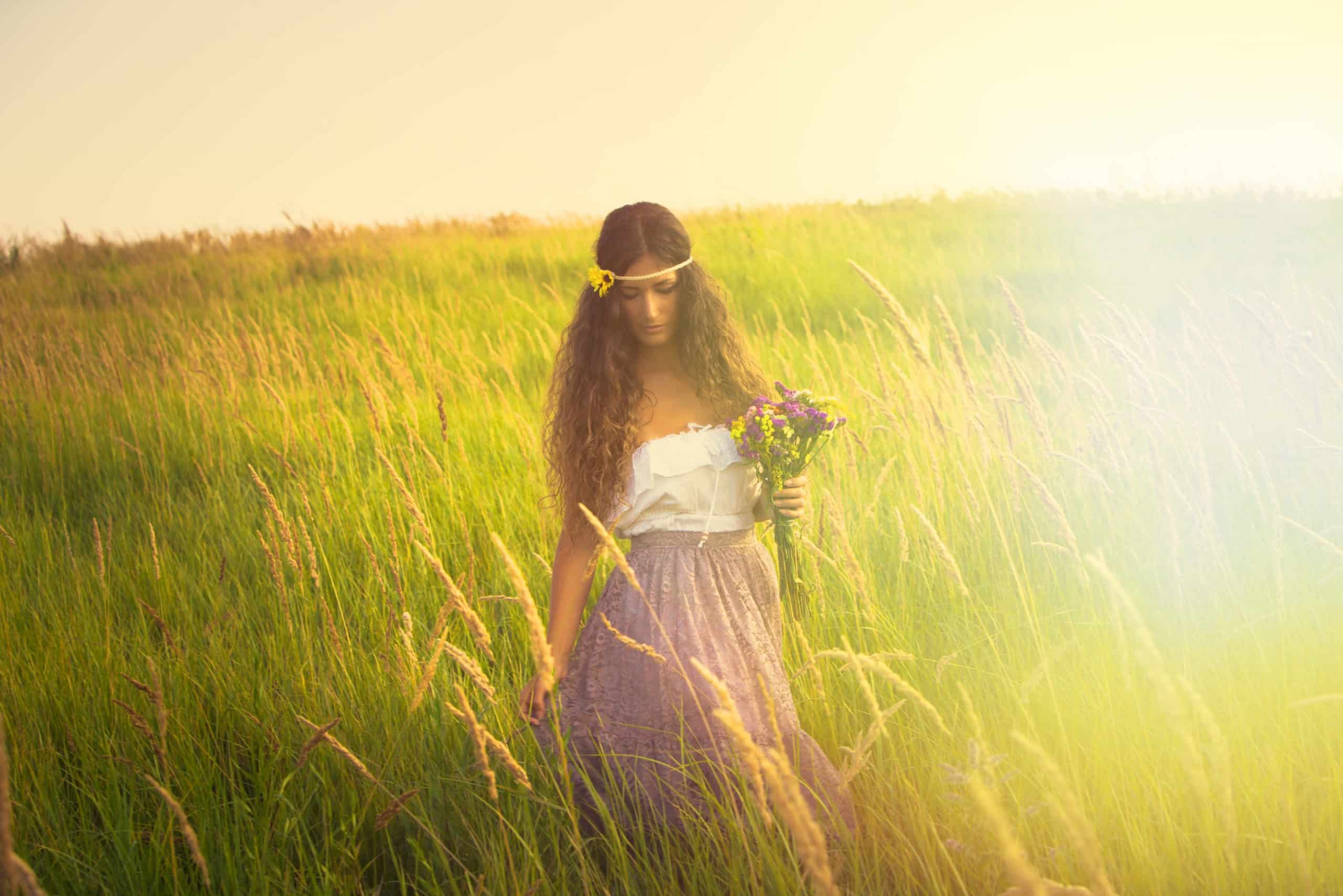 Young beautiful woman with long curly hair hold in hand a bouquet of wild flowers walk in light at grass field