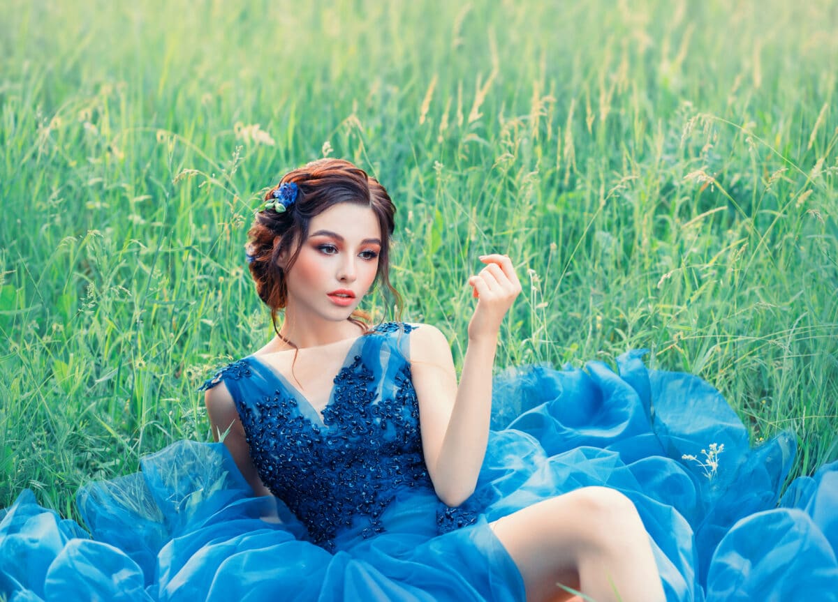 charming mysterious story about porcelain doll, lovely girl in long blue lush delicate dress. lady with dark braided hair and gentle make-up, perfect skin, flower in green grass on fresh nature