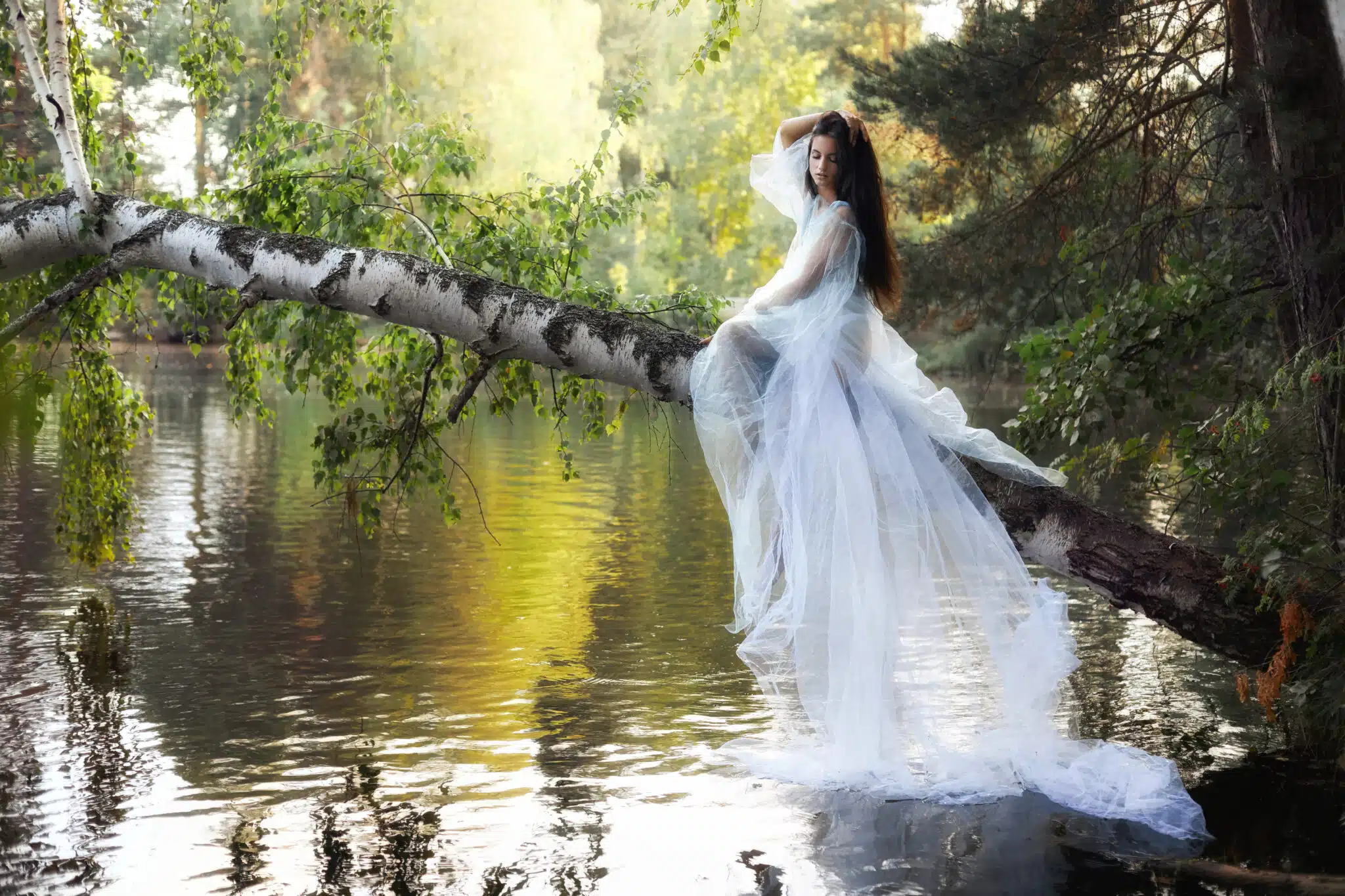 beautiful romantic woman in white long lace dress sitting on fallen tree above river
