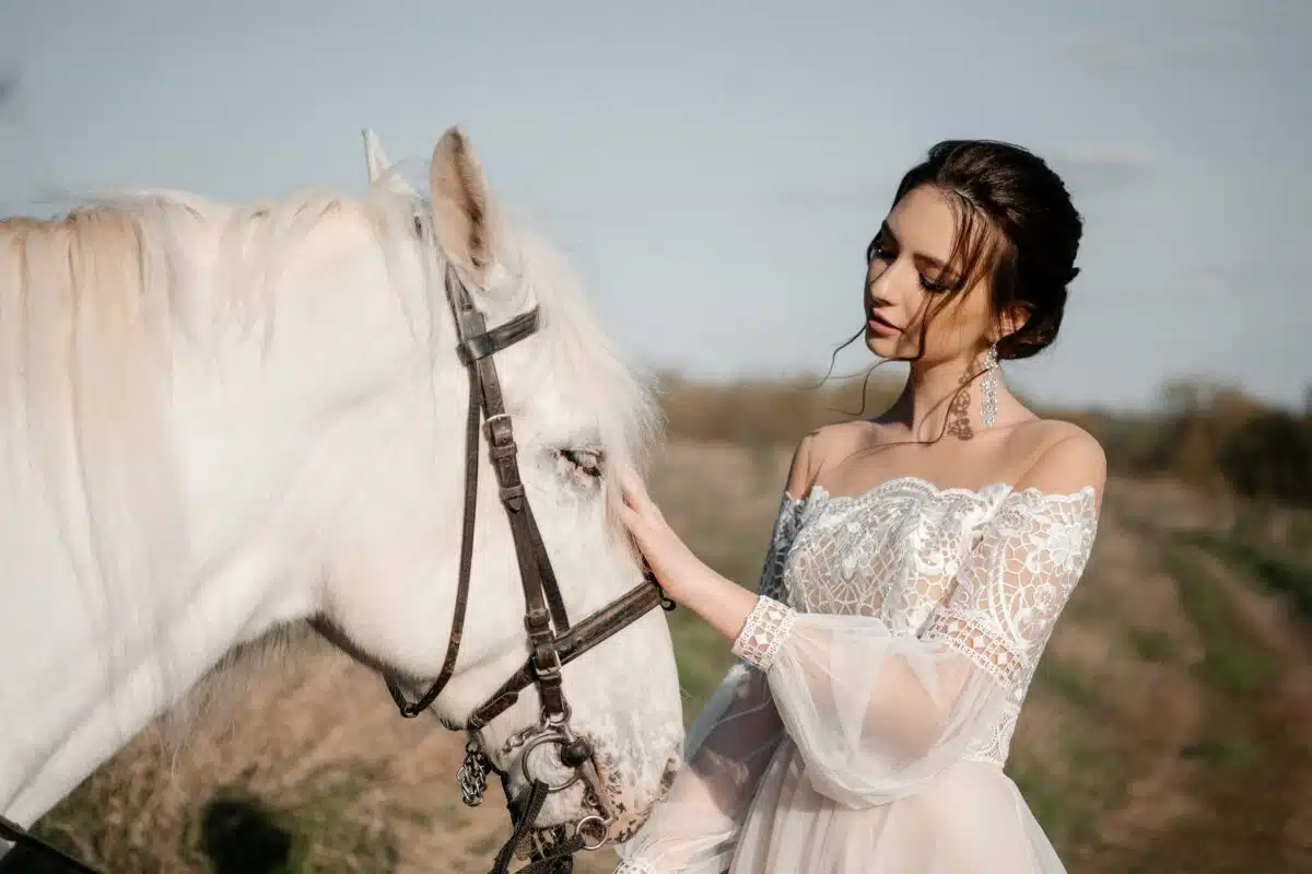 A beautiful lady in white light dress touching her white horse 