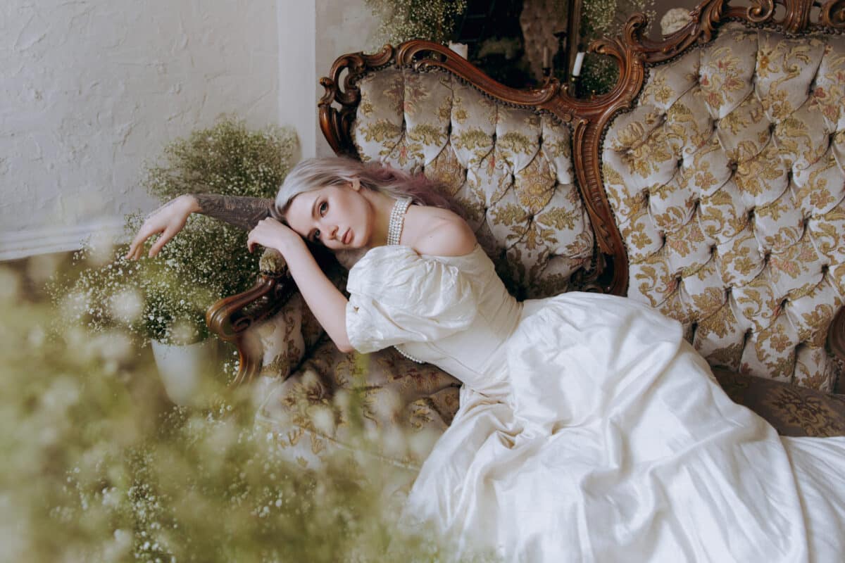 A sad-looking princess in a white dress lying on a luxurious sofa surrounded with white flowers