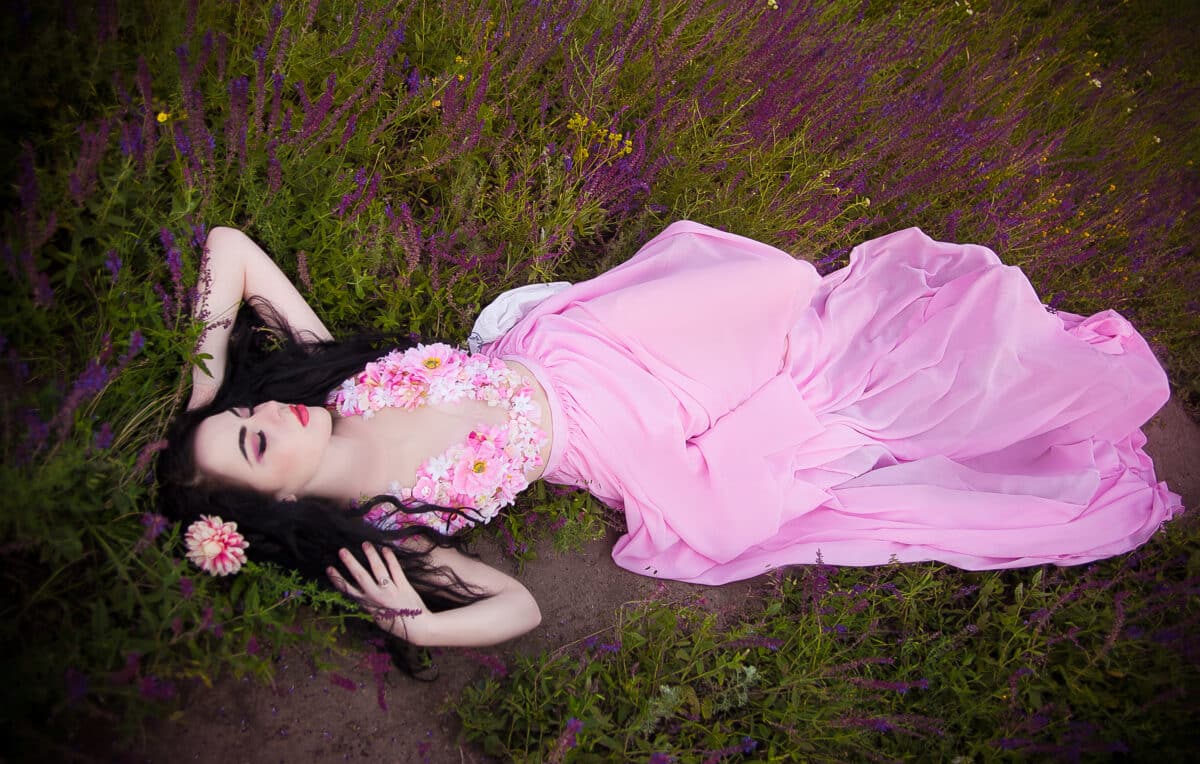 a beautiful brunette woman with long hair in a pink chic floral dress lies on green grass with purple field flowers