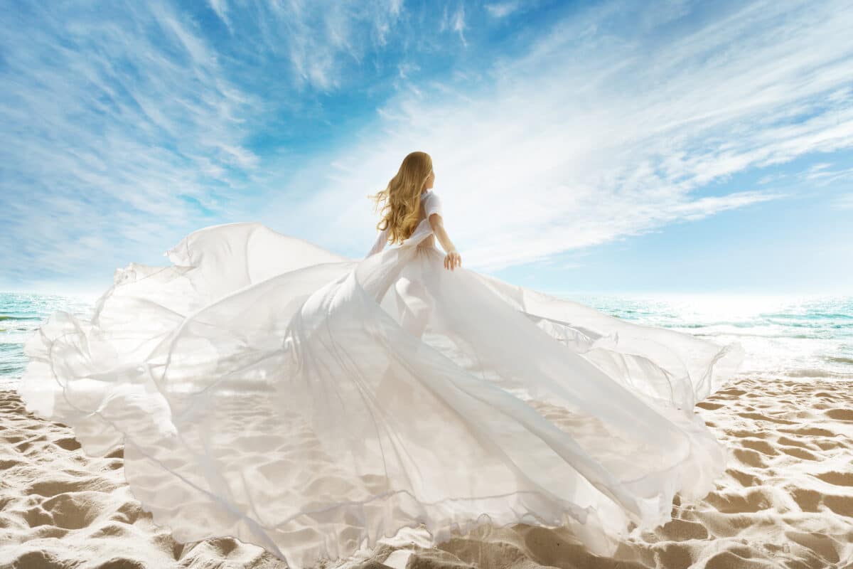 fantasy woman dressed in long white dress walking on the vast sand with the azure sky above her