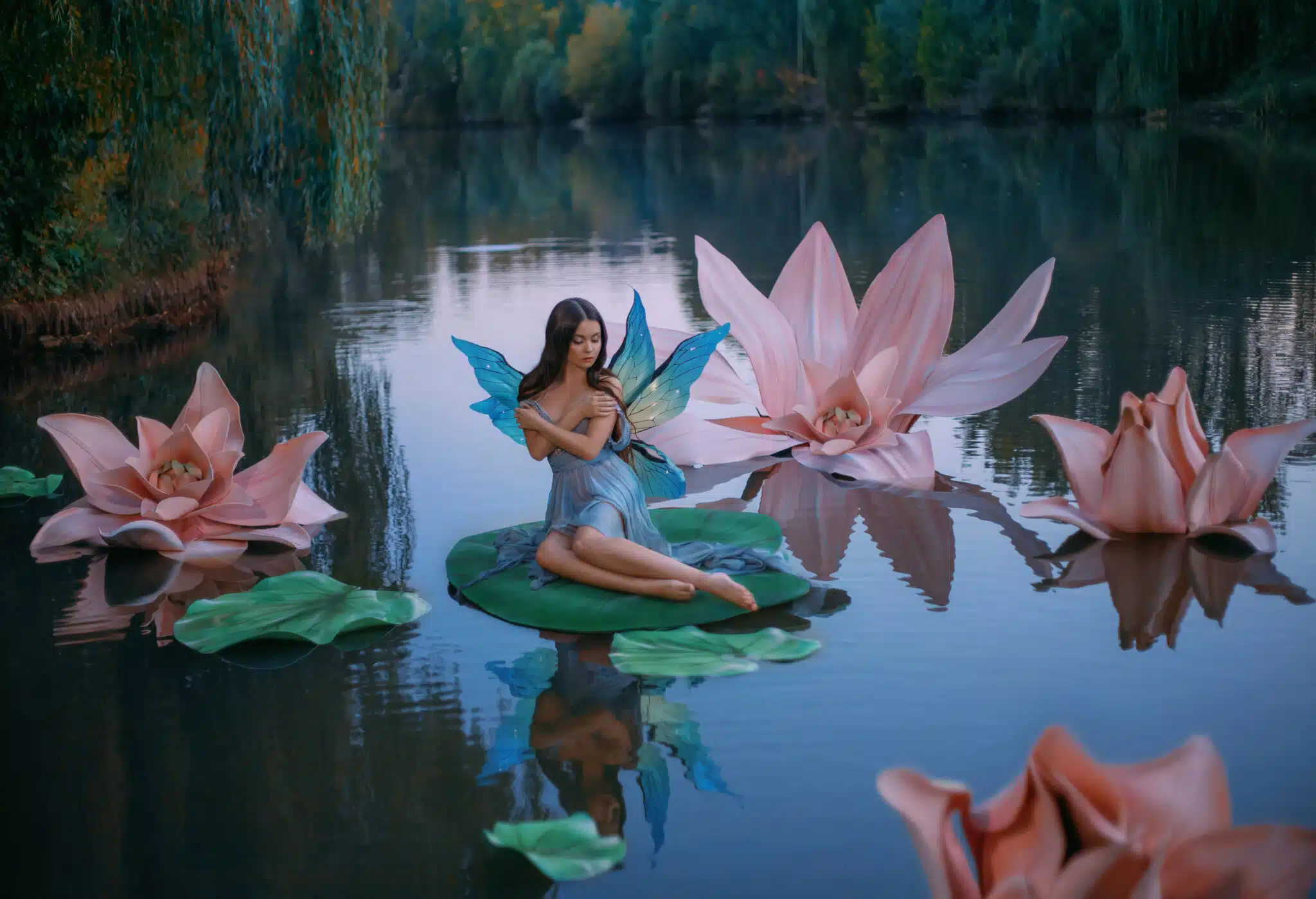 A beautiful woman a little fabulous fairy with butterfly wings sits on green water lily leaf. Fantasy scenery of huge pink flowers on the lake, green trees. River nymph innocent girl in an blue dress
