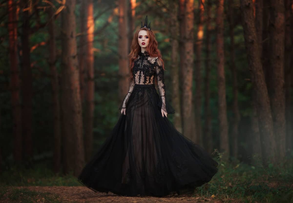 A beautiful gothic princess with pale skin and very long red hair in a black crown and a black long dress in a misty fairy forest. The costume of the dark queen.