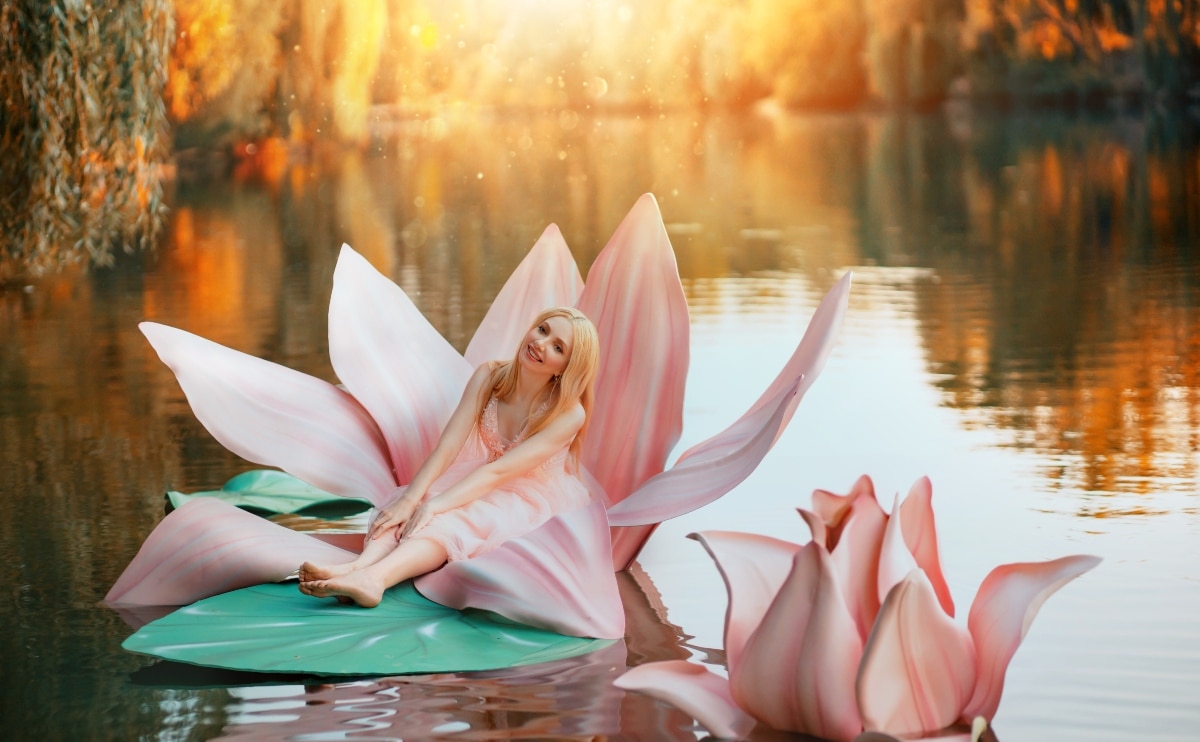 Happy fantasy young blonde woman little fairy princess sitting in pink lotus flower on lake water.