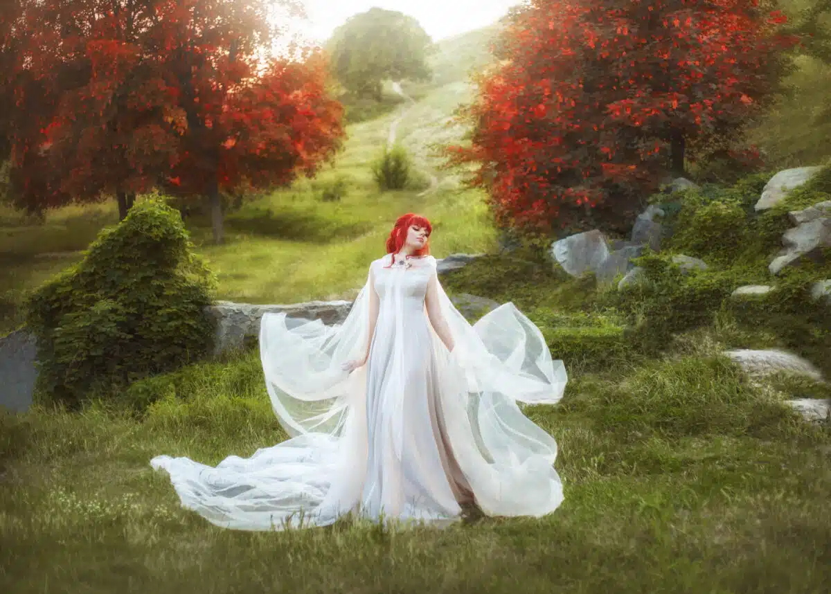 A young, red-haired elf waving a raincoat against the backdrop of autumn hills and a path that leaves at a distance. The wizard, like a bird, flaps her wings from the fabrics, in a white flying dress