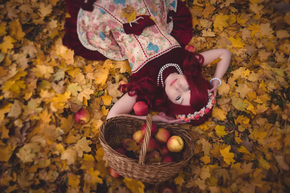 redhead maiden in burgundy vintage dress lies on maple leaves with apples inside the basket