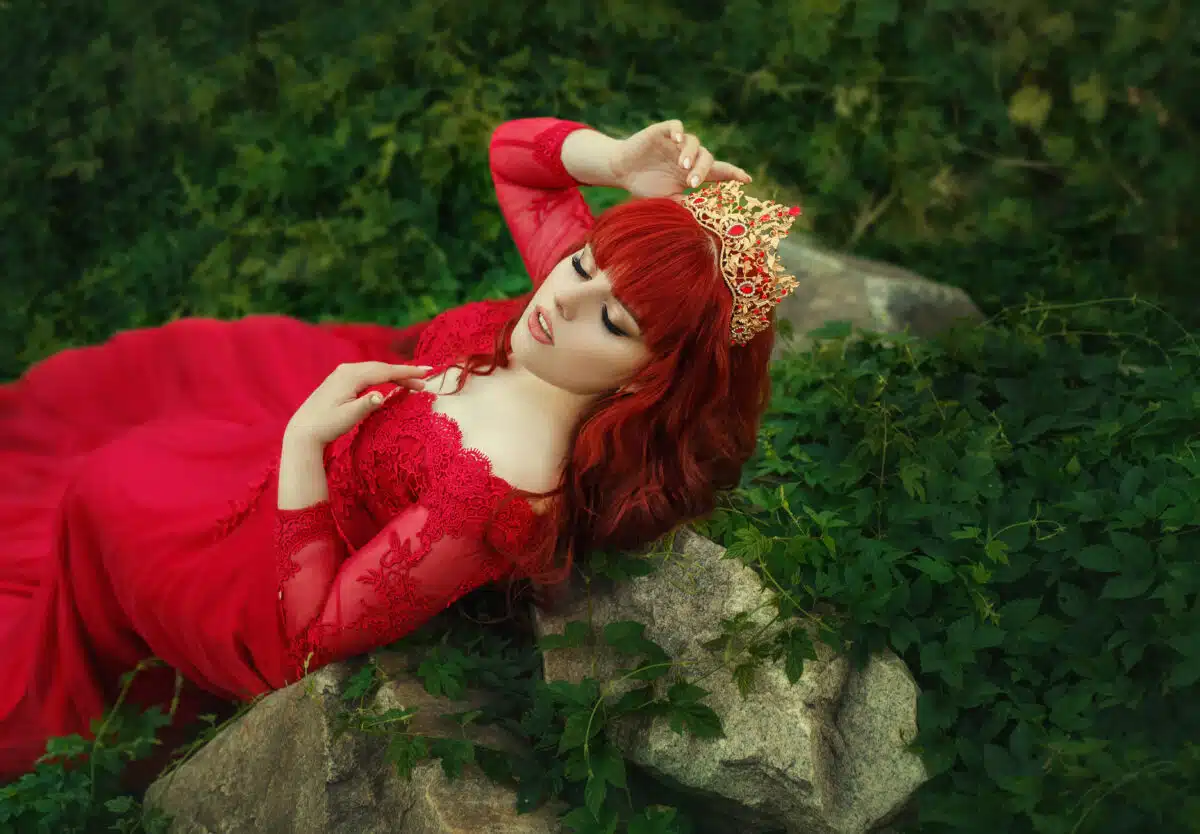  Red-haired woman in a red dress wearing a gold crown while relaxing in nature