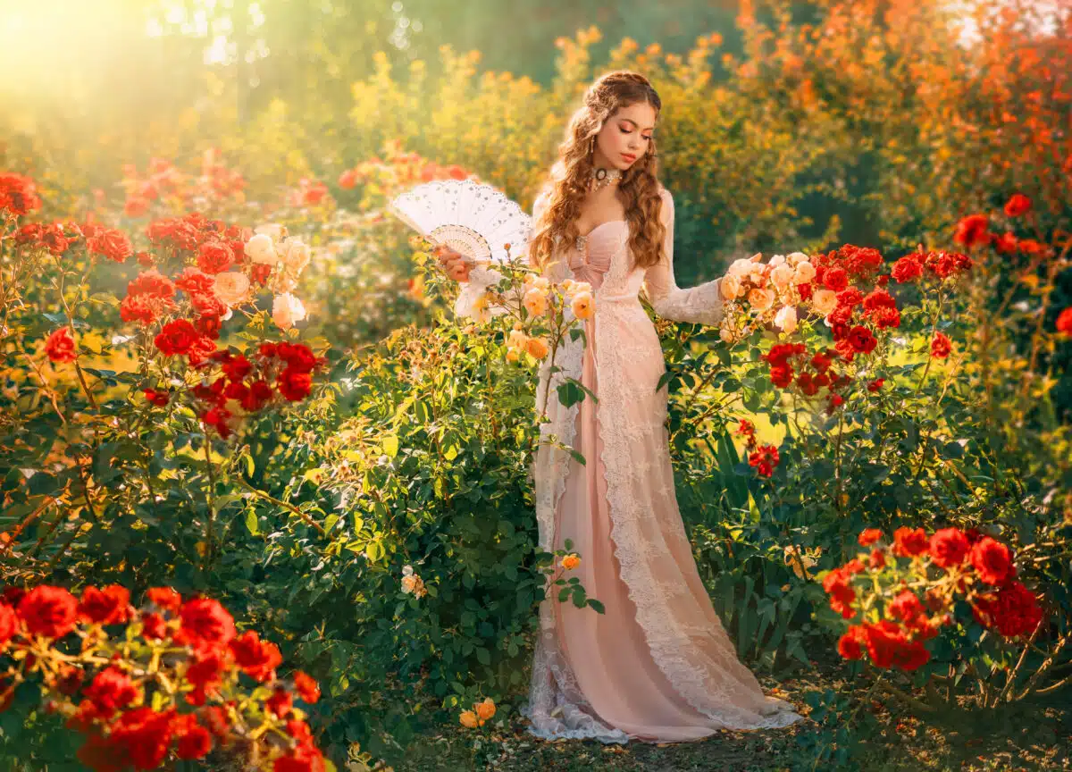 a medieval princess holding fan in hands enjoying in the sunny summer nature garden with red roses 