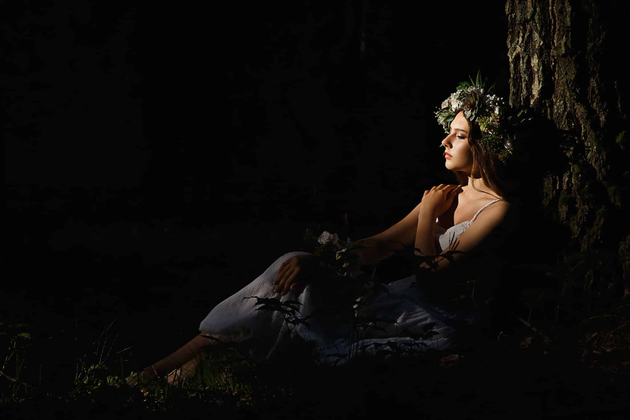 a mysterious woman in gown with flower wreath sitting in dark room