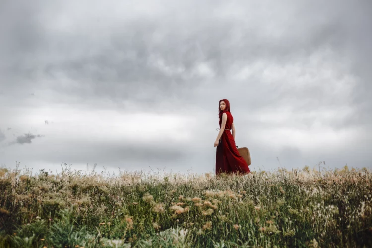 A lonely red hair girl in red dress holding a vintage suitcase walking in nature