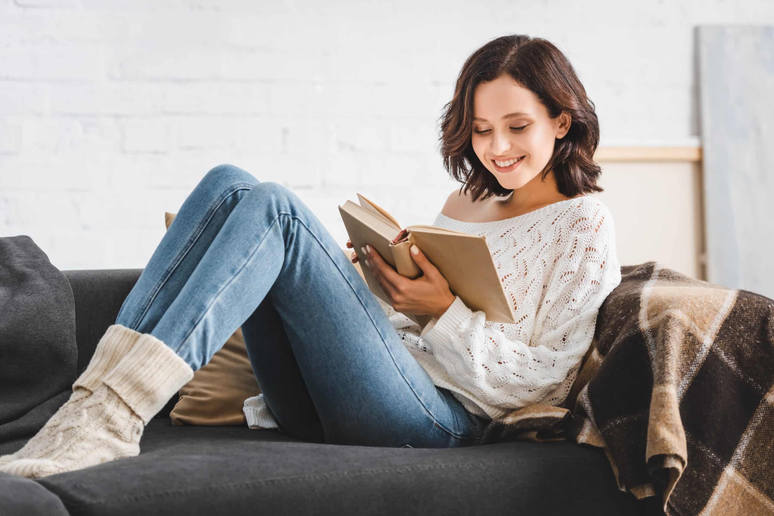 Smiling young woman reading book on sofa at home.