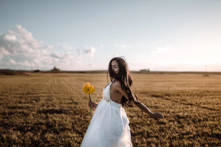 Young Hispanic female in a beautiful white dress holding a flower and running in a field at sunset