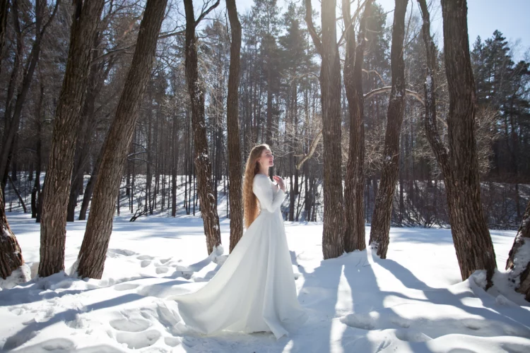 A young blonde woman in a long white dress walks in the snow forest