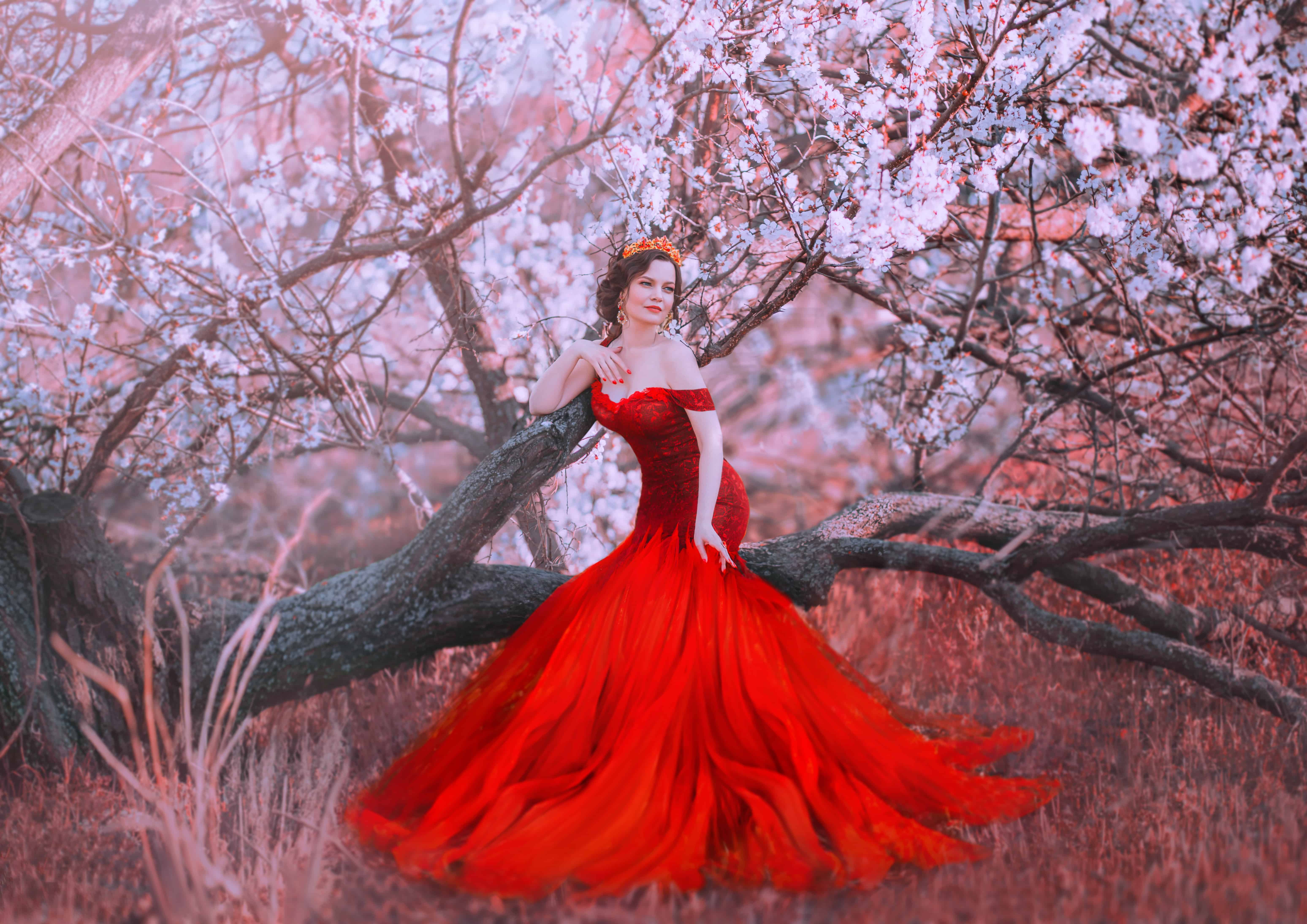 charming nymph sits on fallen tree in spring forest, lady in gorgeous red scarlet long dress with bare shoulders, mermaid becomes human , fairy-tale princess walks in sakura garden, creative colors
