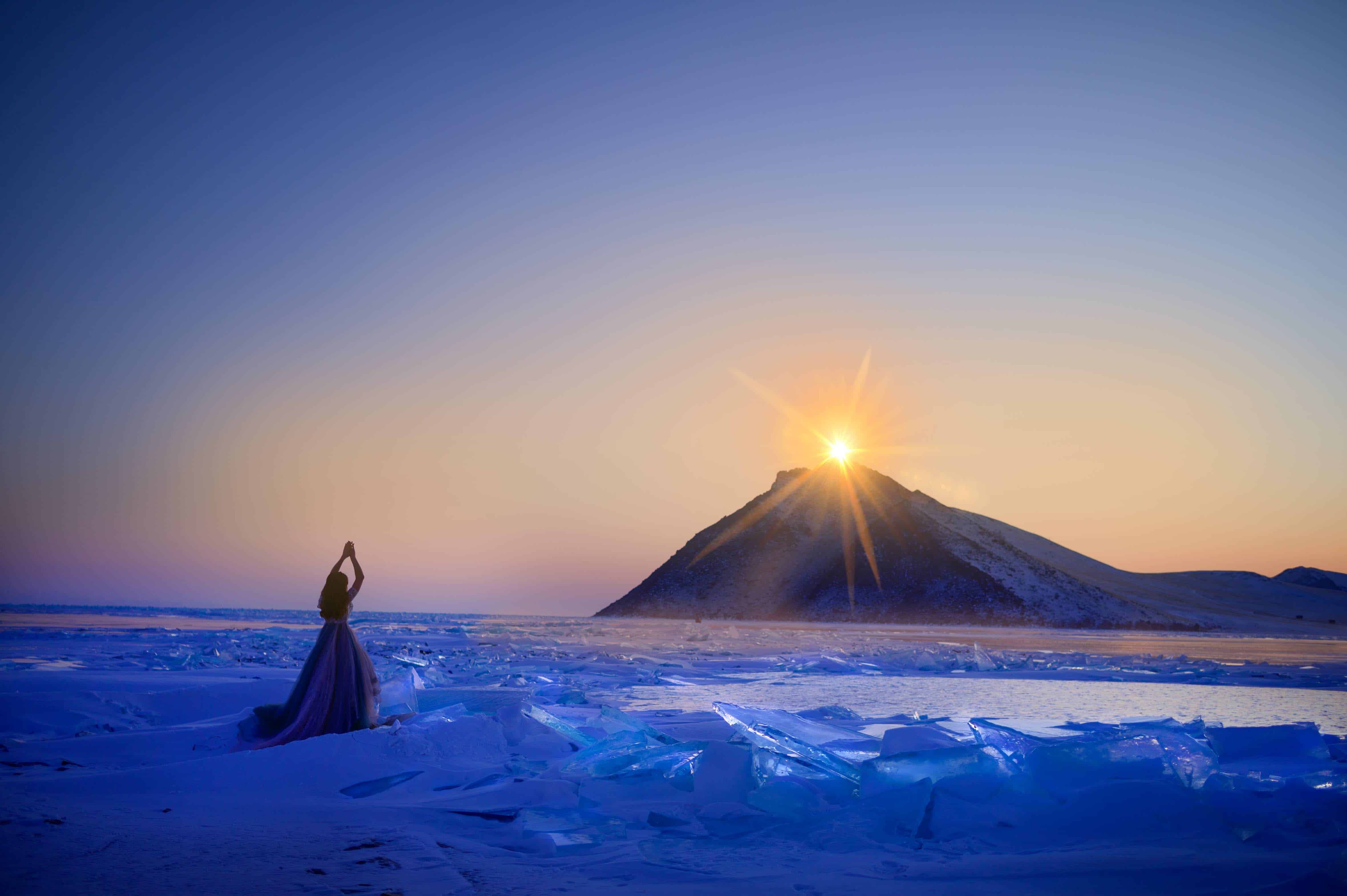 mysterious woman in a blue fluffy dress raising hands in prayer looking at the rising sun on snowy mountain