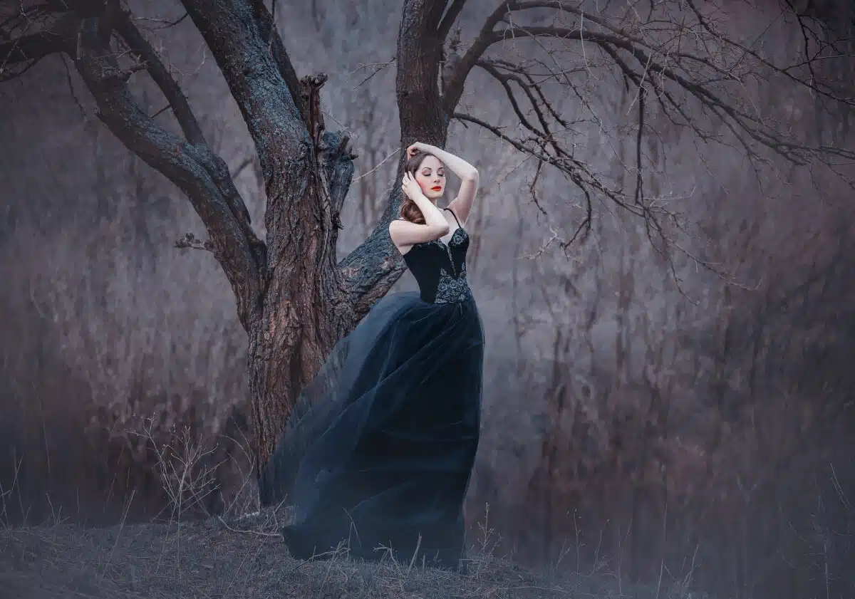 enchanting lady in a long black dress with bare open arms and shoulders standing alone in the autumn cold forest
