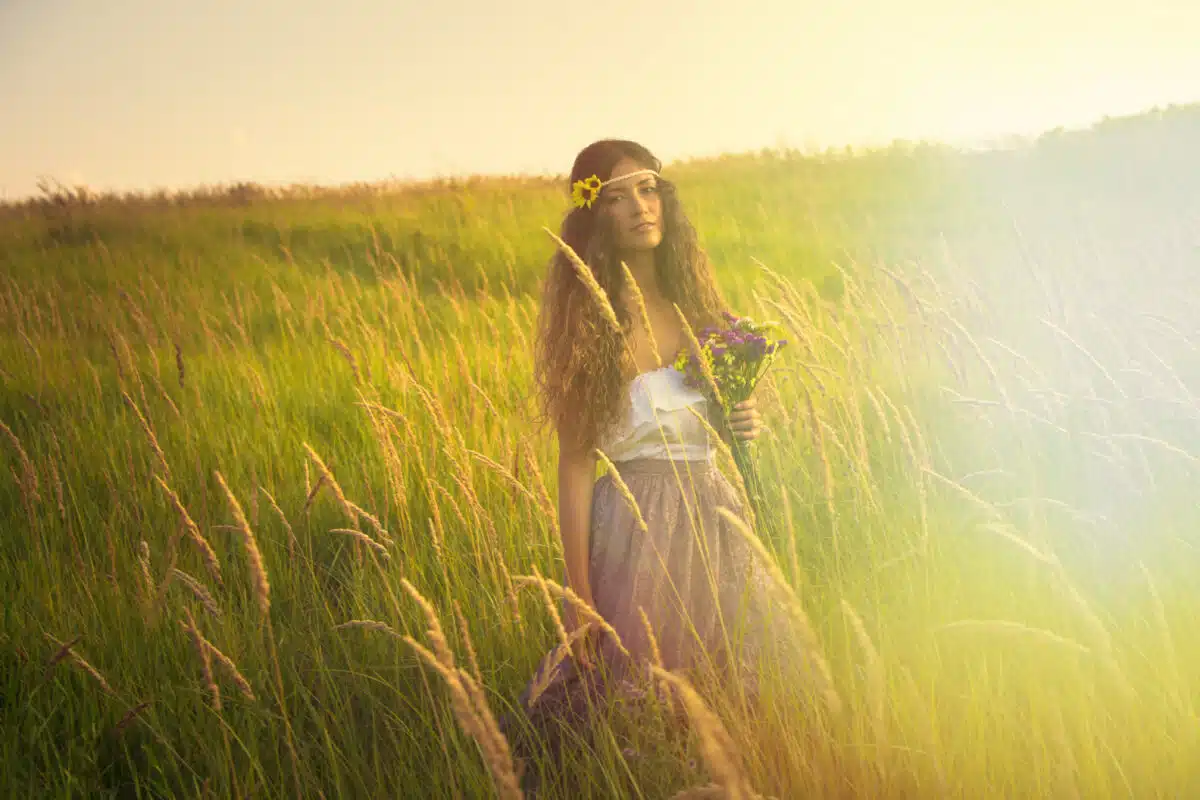 a young beautiful woman with long curly hair holding a bouquet of wild flowers in romantic clothes while walking in grass field at daylight