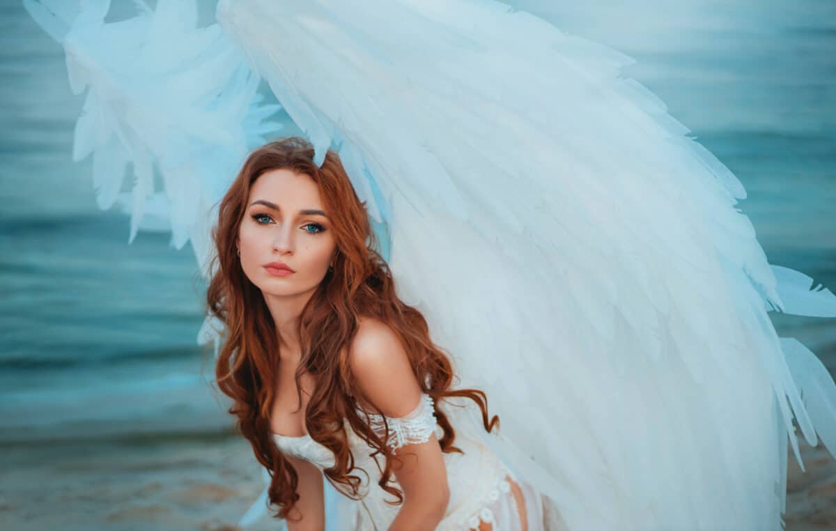 Portrait of an attractive adult angel girl, innocent face blue eyes. Beautiful fashion model young fantasy Woman with white bird wings feathers. Greek goddess costume. Background - nature, water river